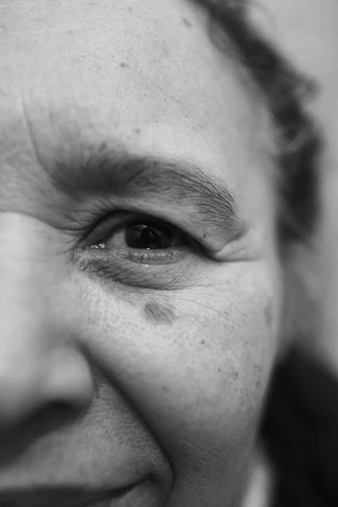 Close-up black and white photo of a smiling person's face, focusing on the eye and cheek.
