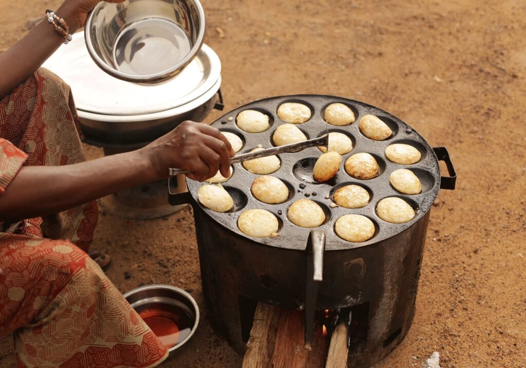 Person cooking small round dough balls on a griddle over an open fire outdoors in a traditional setting.