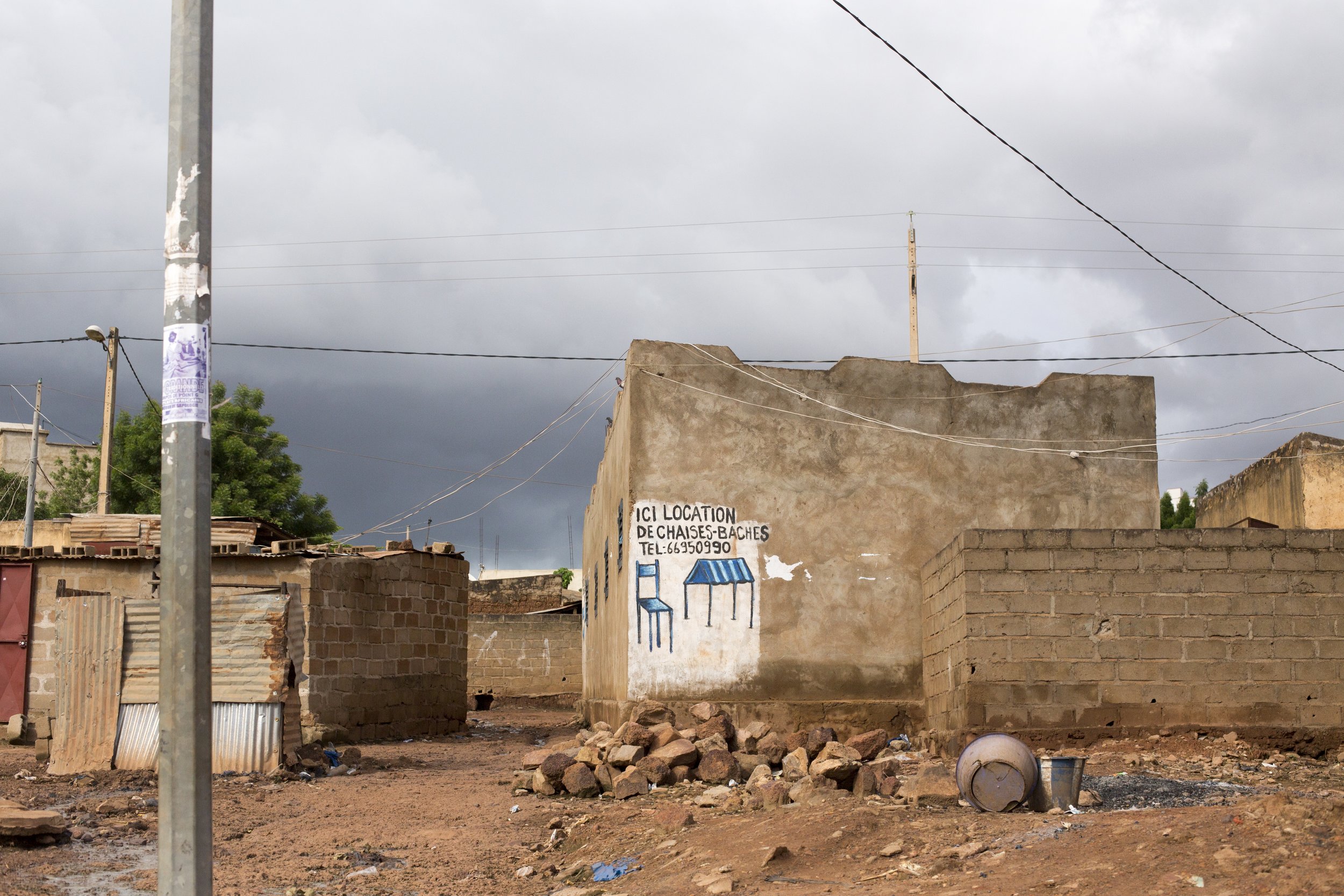 A weathered wall in a dirt area with a painted sign advertising chairs and benches, including a phone number, and a stormy sky overhead.