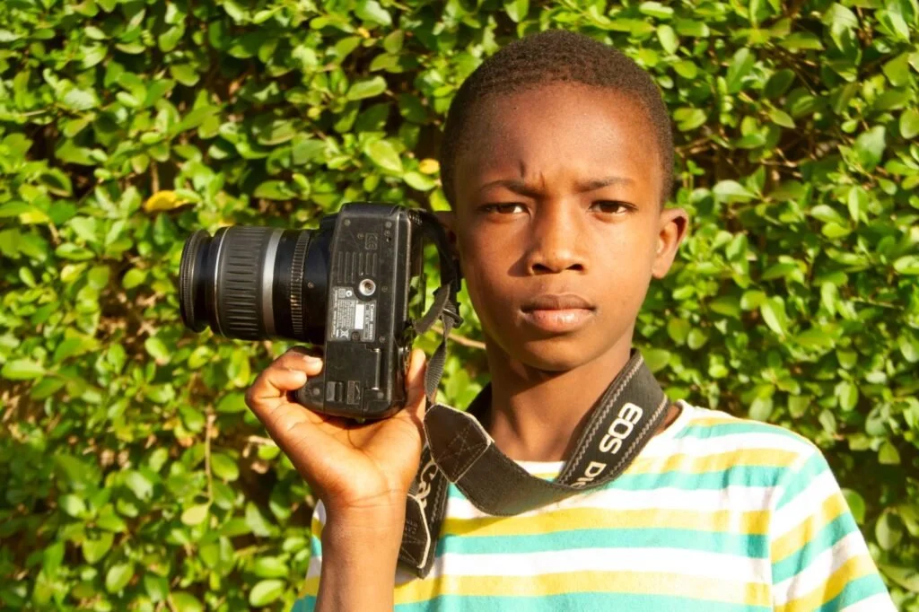 A young boy holding a DSLR camera with a strap around his neck, standing in front of green foliage.