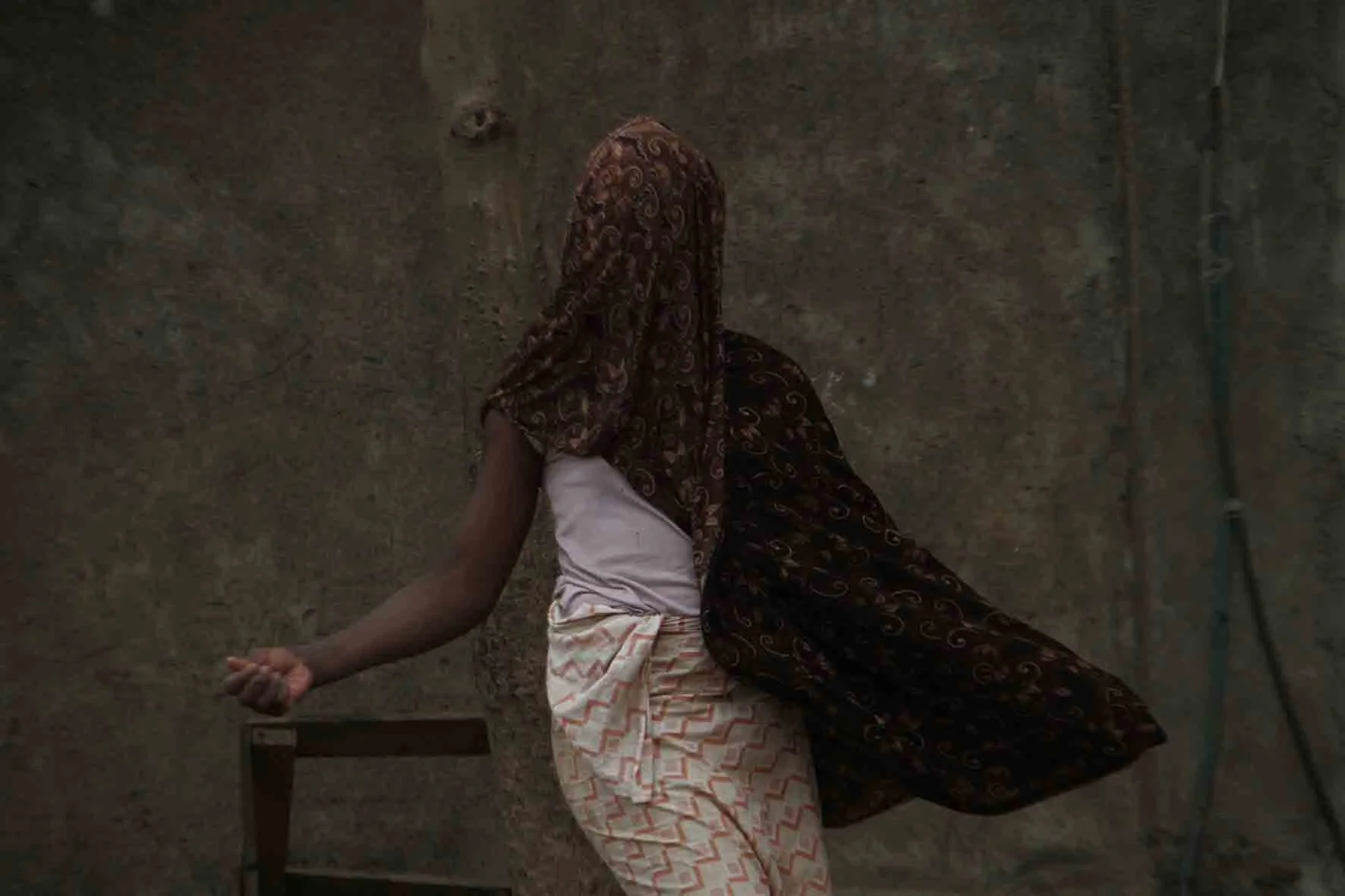 A woman is standing with her back to the camera against a textured wall, wearing a patterned head covering, a white sleeveless top, and patterned pants, with her right arm extended and her hand open.