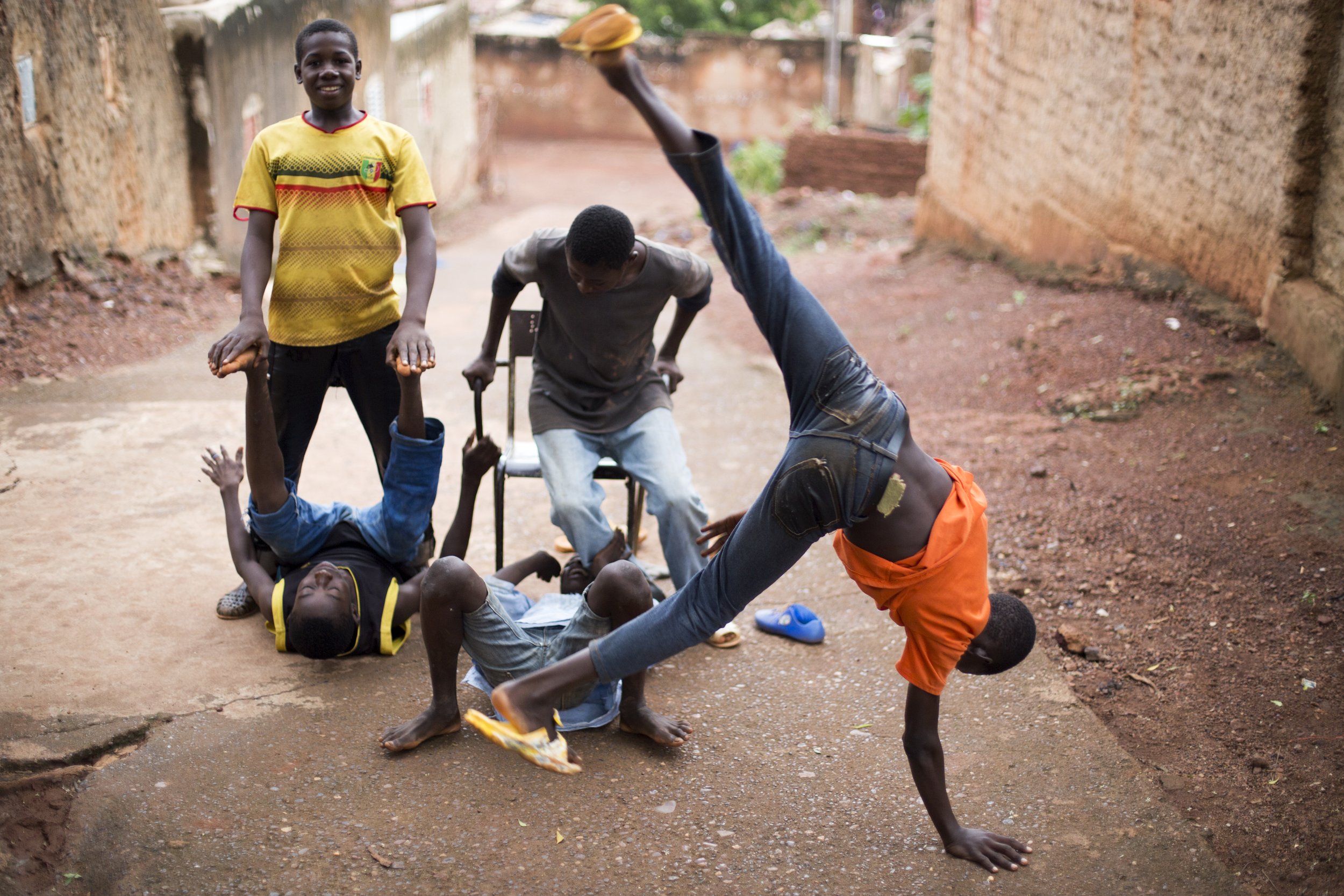 Group of children playing on a dirt path, with one boy performing a handstand, two boys lying on the ground, one standing, and another with a crutch.