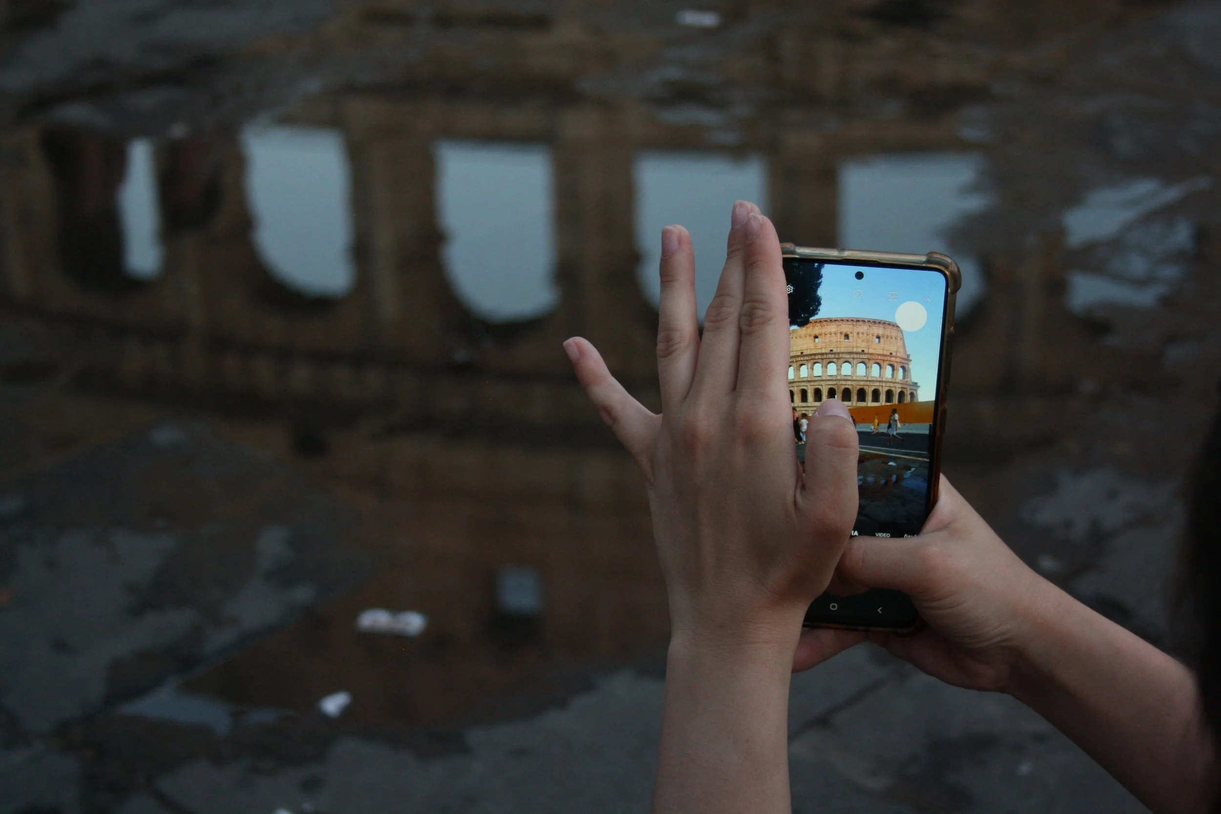 Person taking a photo of the Roman Colosseum with a smartphone, with reflections in water on the ground.