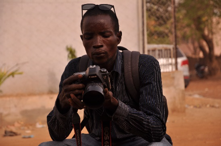 A man with sunglasses on his head, wearing a dark shirt, sitting outdoors and looking at a camera in his hands with a backpack on his shoulder.