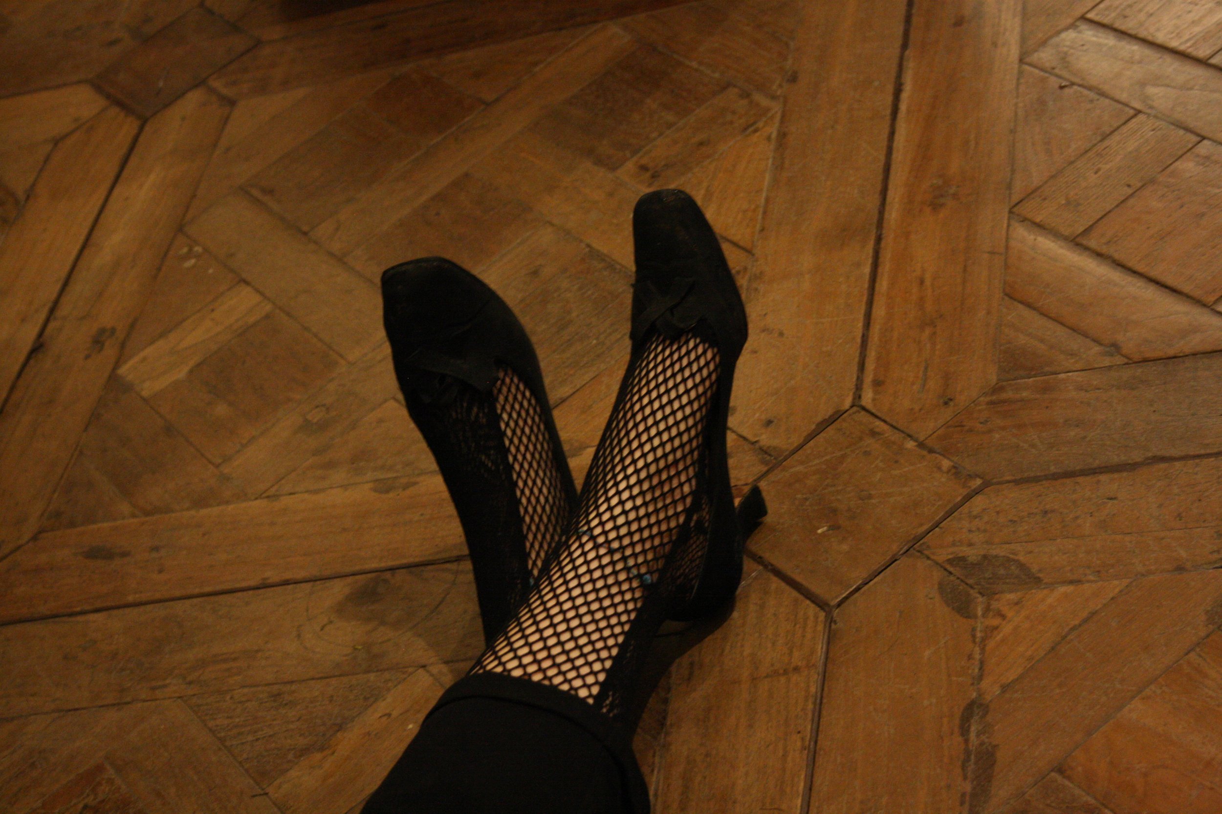A person's legs wearing fishnet stockings and black shoes, standing on a wooden floor.