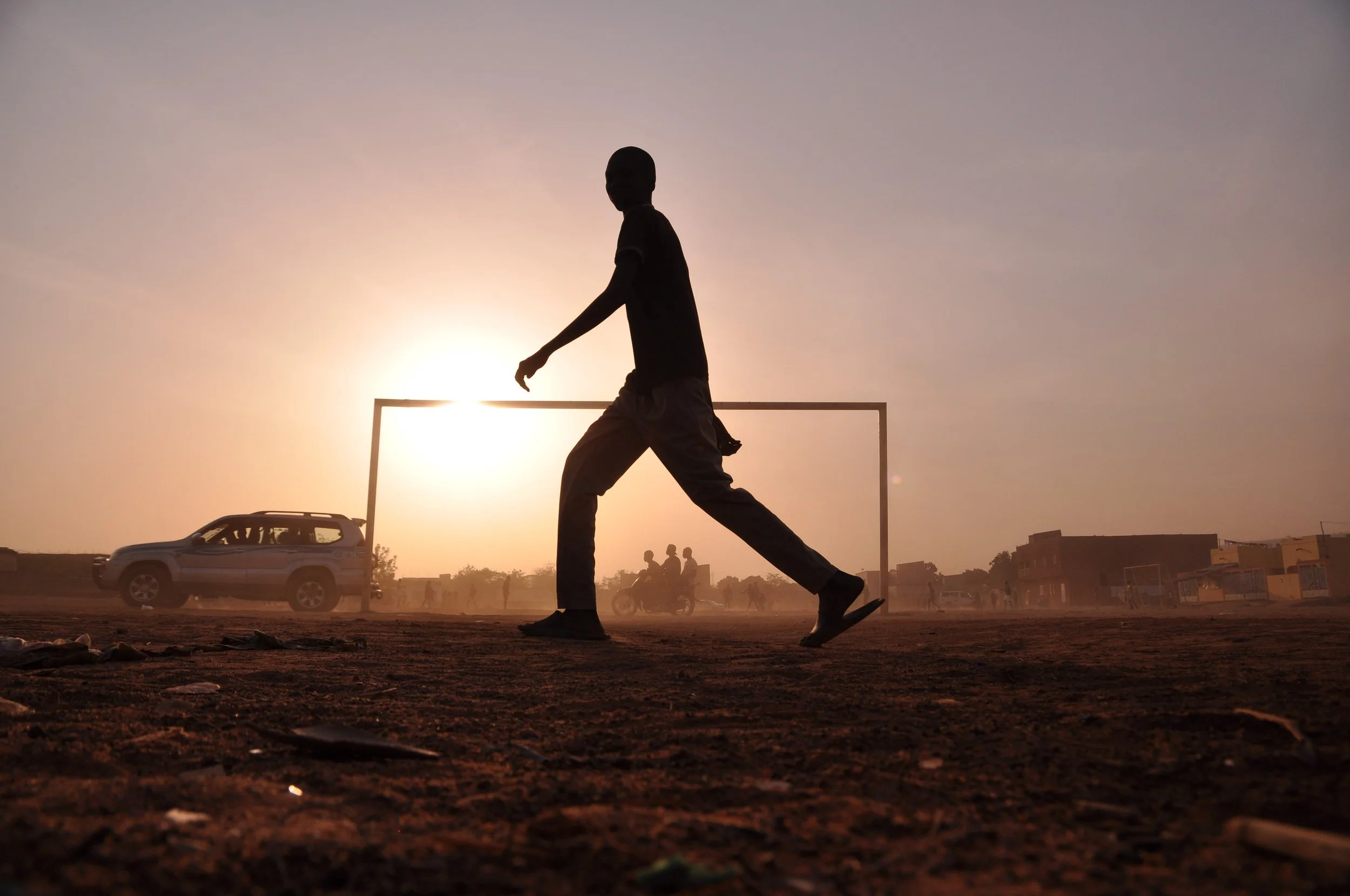 Silhouette of a boy playing soccer at sunset on a dirt field, with a goalpost, cars, and buildings in the background.