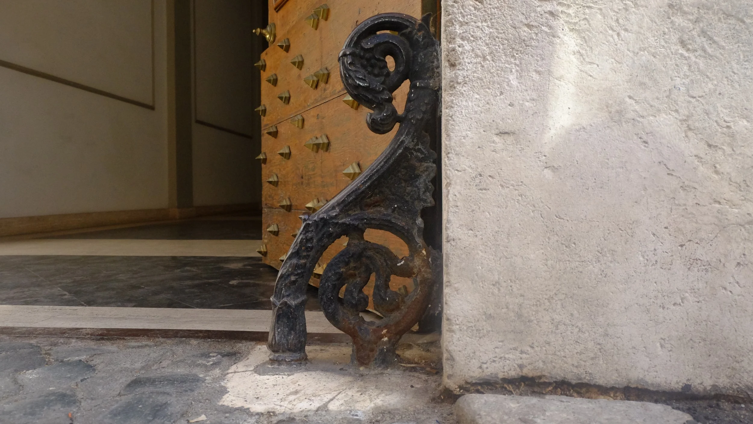 Close-up of an ornate black cast-iron doorstop shaped like a seahorse with a curled tail, placed on a stone floor near a concrete wall, with a wooden piece of furniture with metal studs in the background.