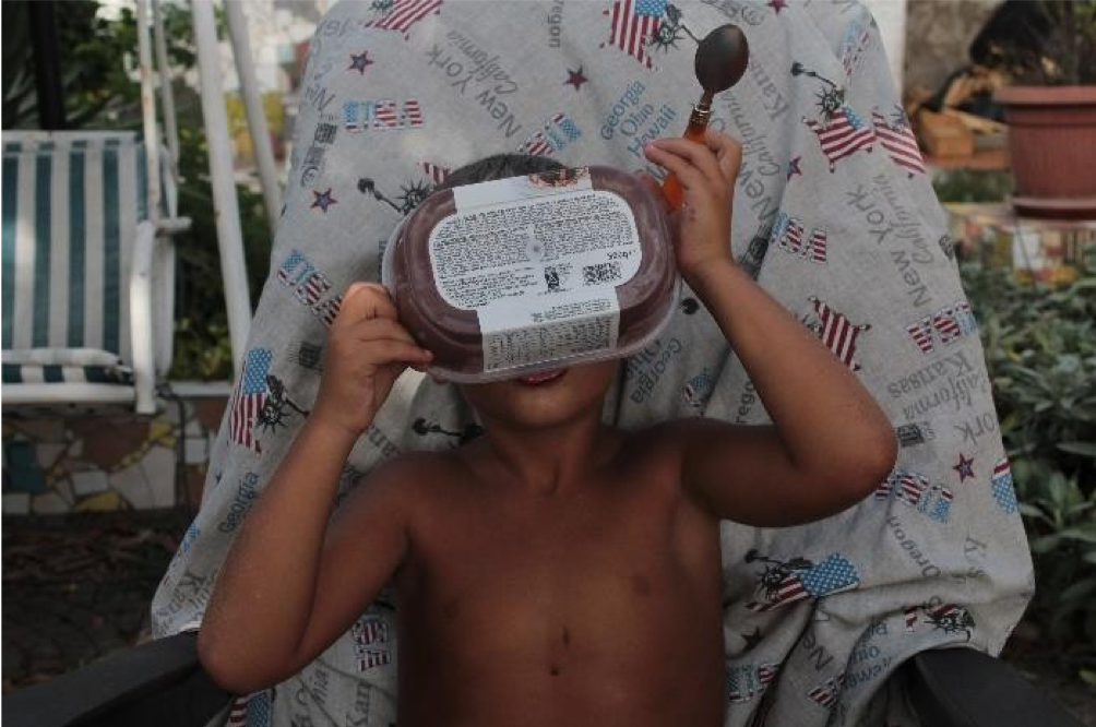 A young boy sitting outdoors in a chair with a patriotic themed towel, holding a container of food to his face as if it were a mask, with a spoon in his other hand.