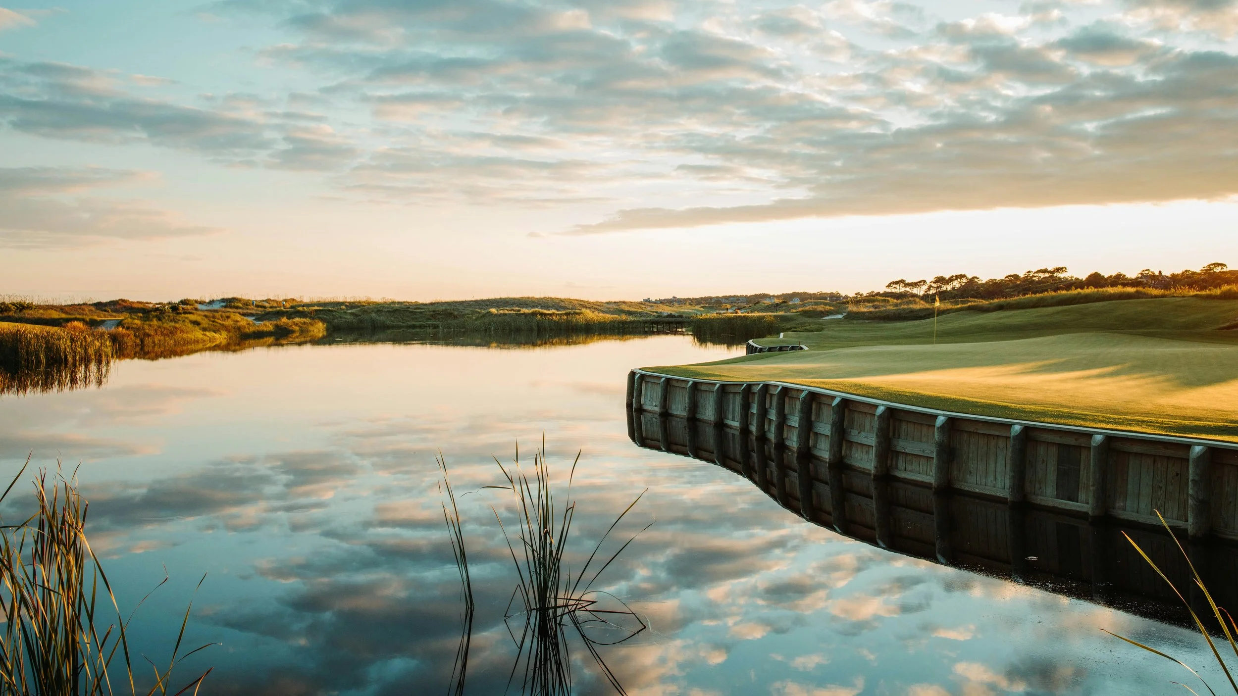 A serene golf course with a water hazard reflecting the cloudy sky during sunset.