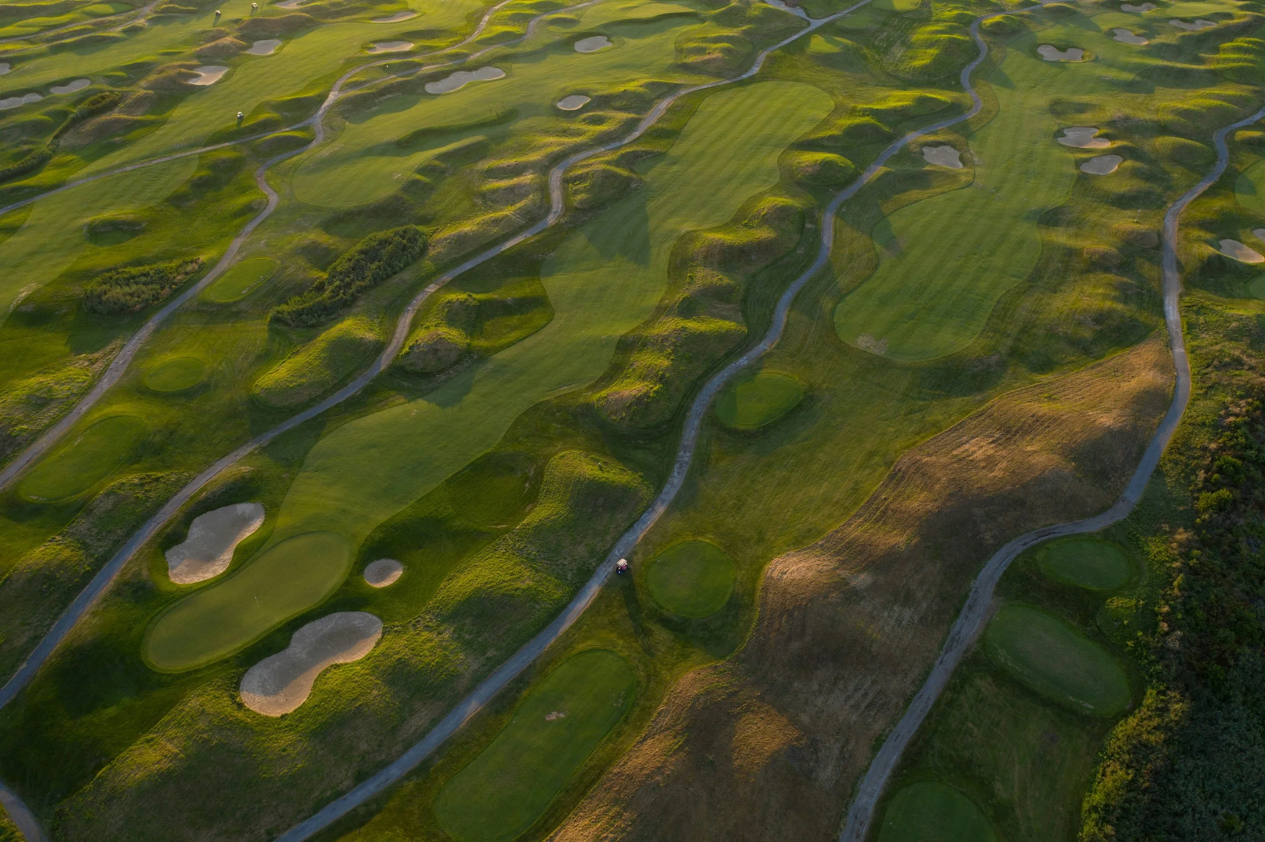 Aerial view of a golf course with multiple greens, sand traps, and winding pathways, surrounded by lush green grass and some trees at the edges.