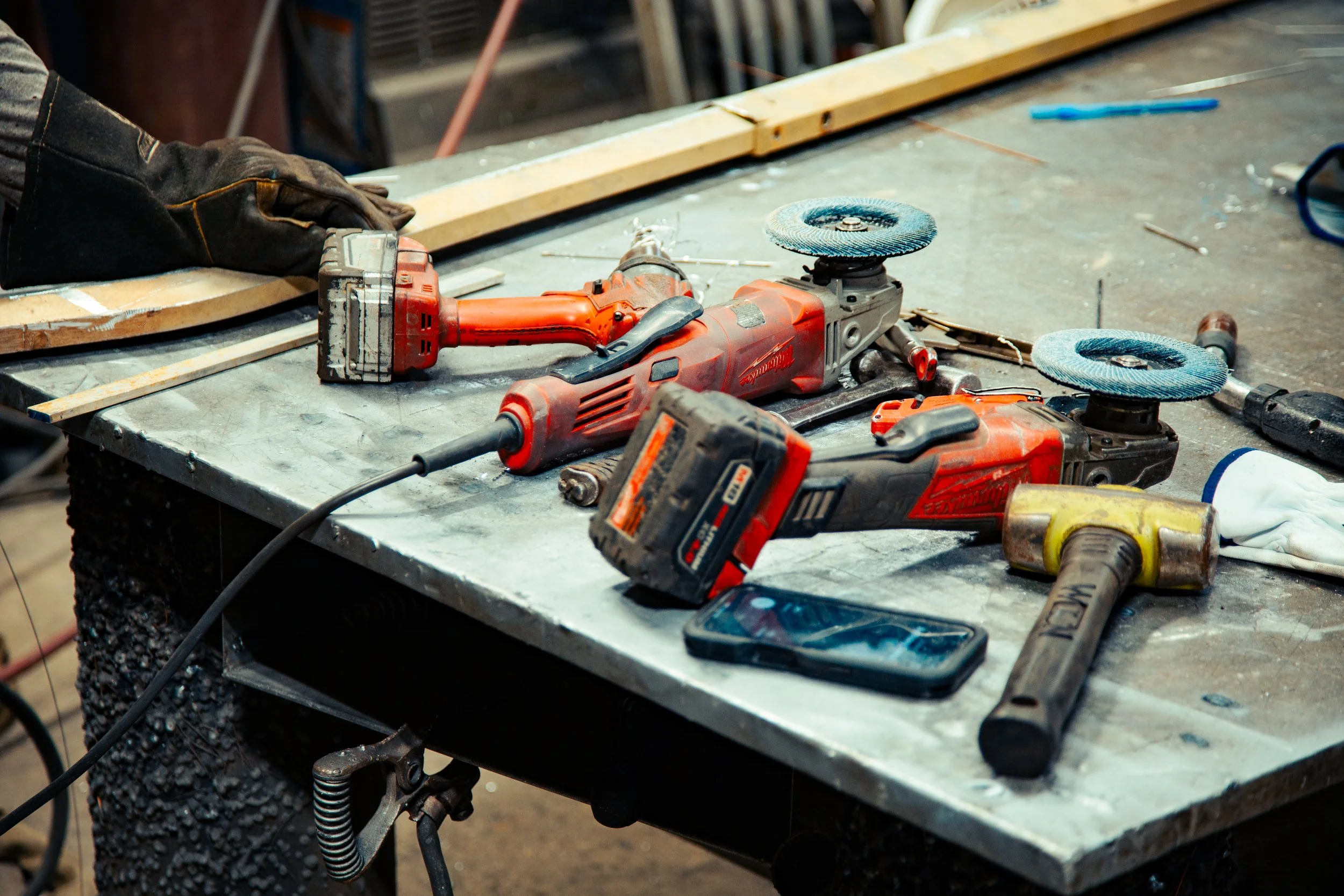 A cluttered workbench with power tools including a cordless drill, an angle grinder, and a rotary hammer, as well as a smartphone, gloves, and small hand tools.
