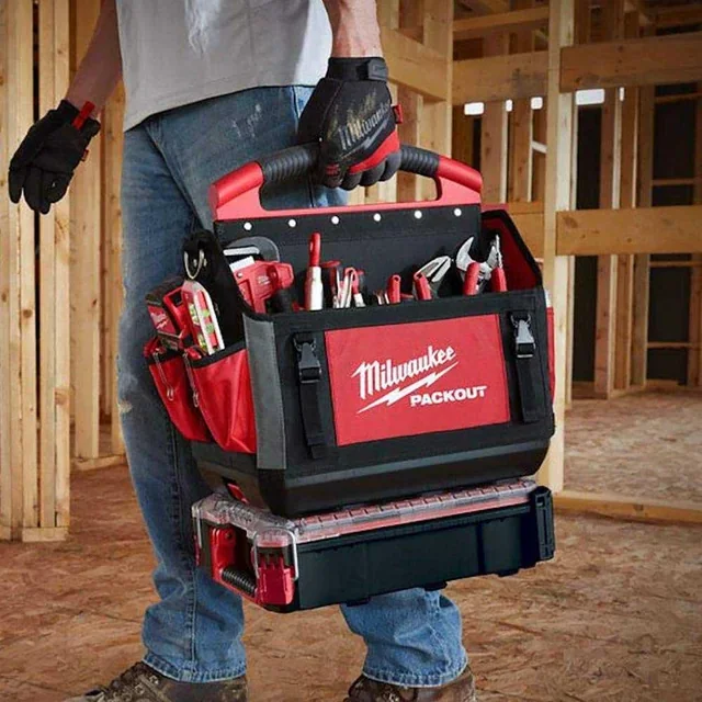 Man holding a Milwaukee Packout toolbox with power tools and hand tools inside, construction site background.