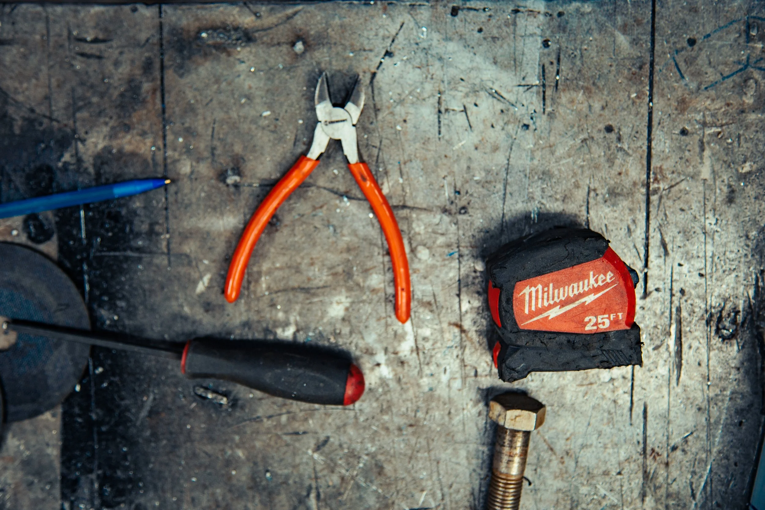 Tools on a worn wooden surface including needle nose pliers, a screwdriver, a measuring tape, a bolt, and a screw.