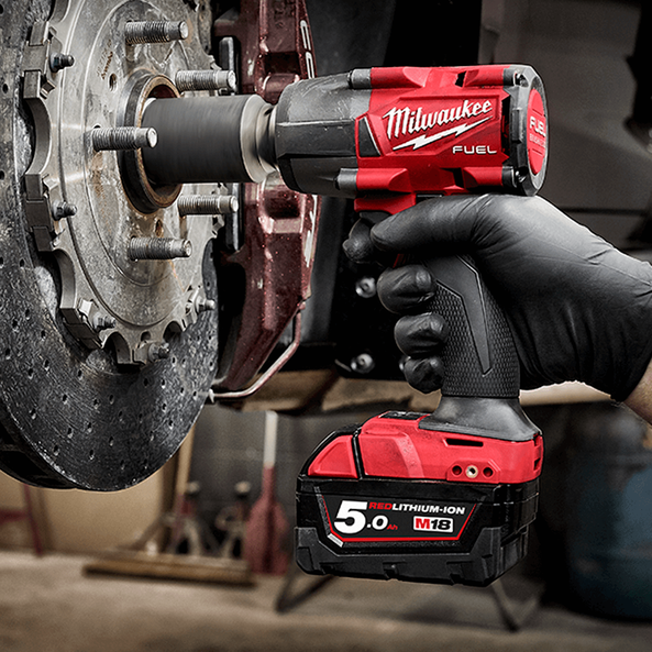 A person wearing a black glove holding a red Milwaukee cordless power tool connected to the back of a car, working on a wheel assembly.