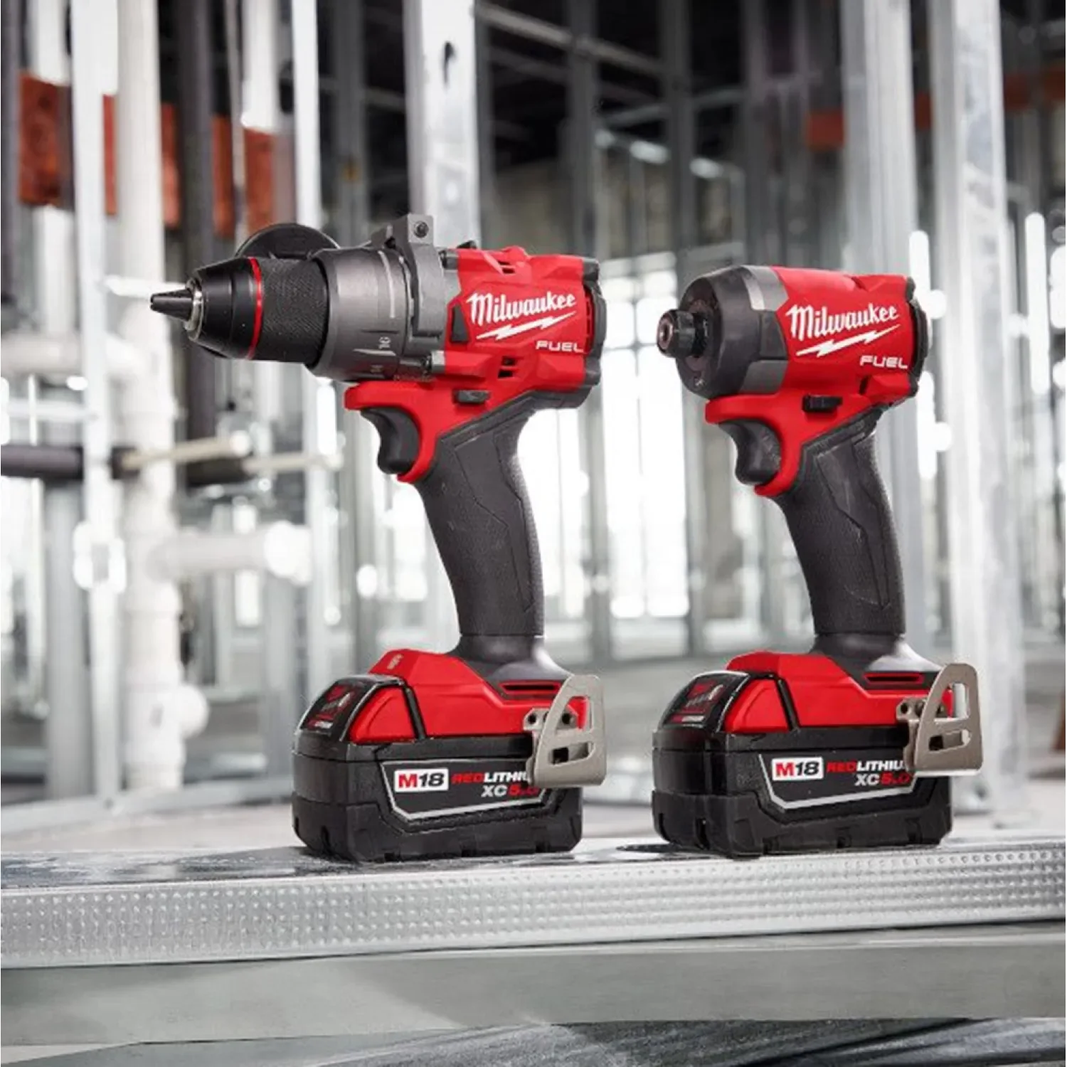 Two Milwaukee cordless power tools, a drill and an impact driver, are placed on a workbench in a construction setting.