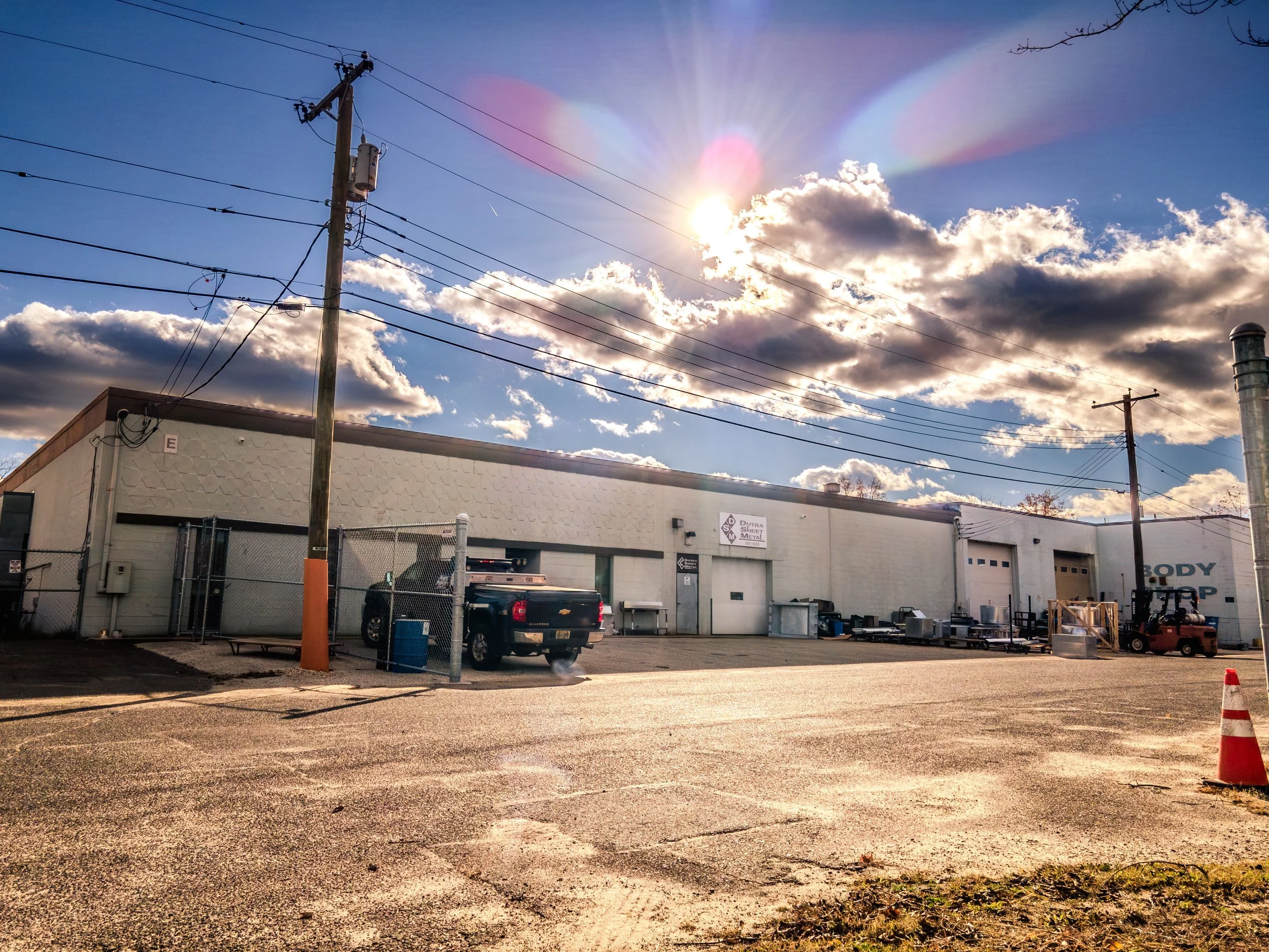 An industrial warehouse with a fenced area, trucks, equipment, and a parking lot under a sky with clouds and the sun.
