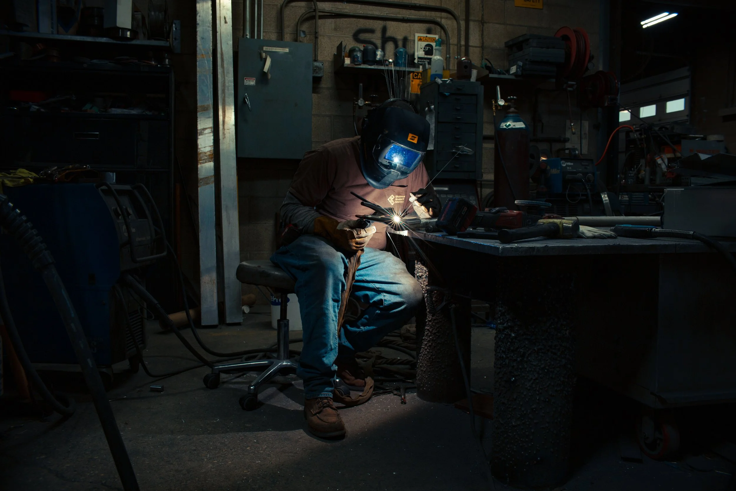 A person welding in a workshop, wearing protective gear including a welding mask, gloves, and casual clothes, with sparks flying from the welding process.