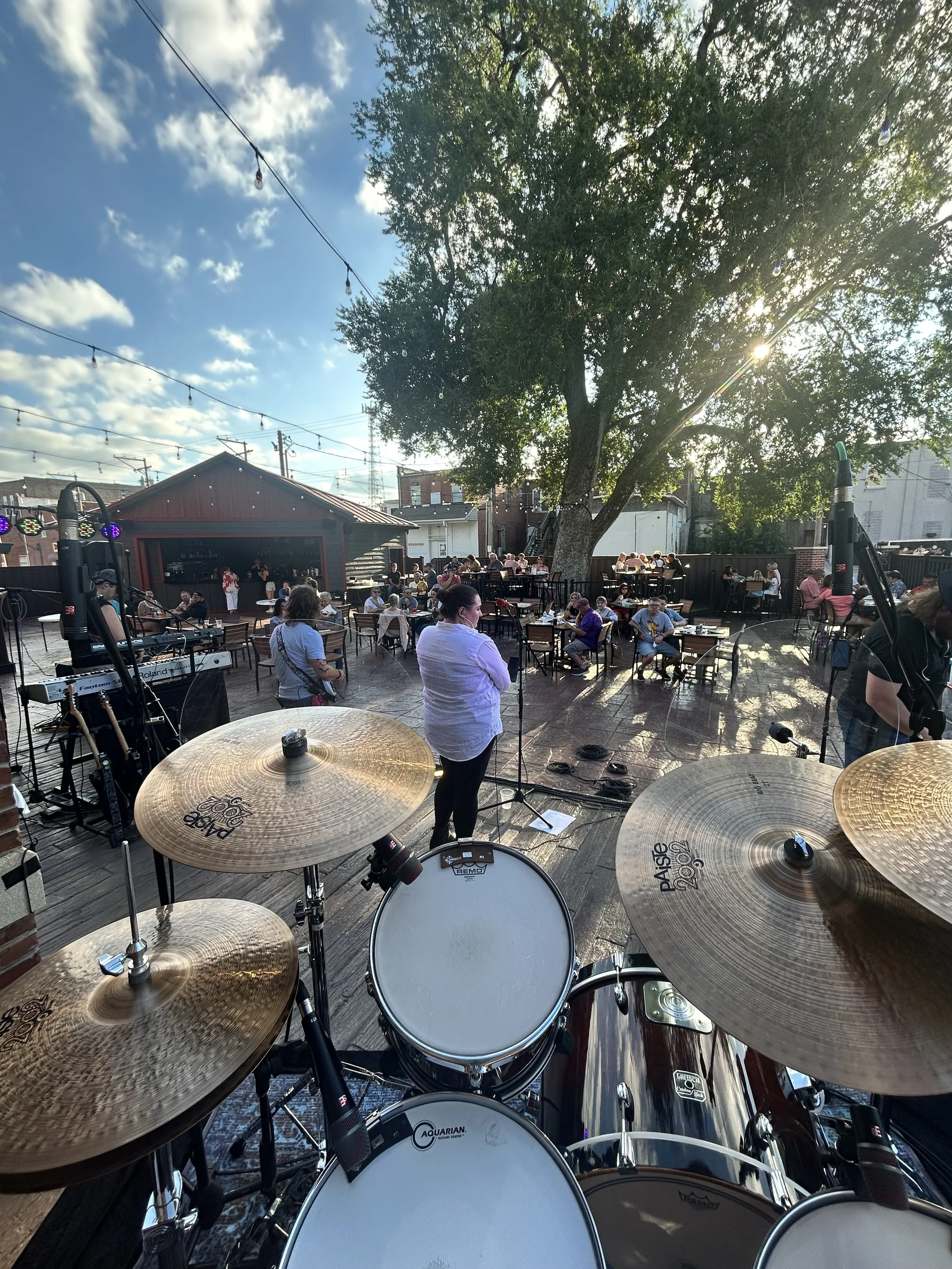View from a drum set looking out onto an outdoor concert at sunset with people seated at tables, a large tree, and string lights overhead.