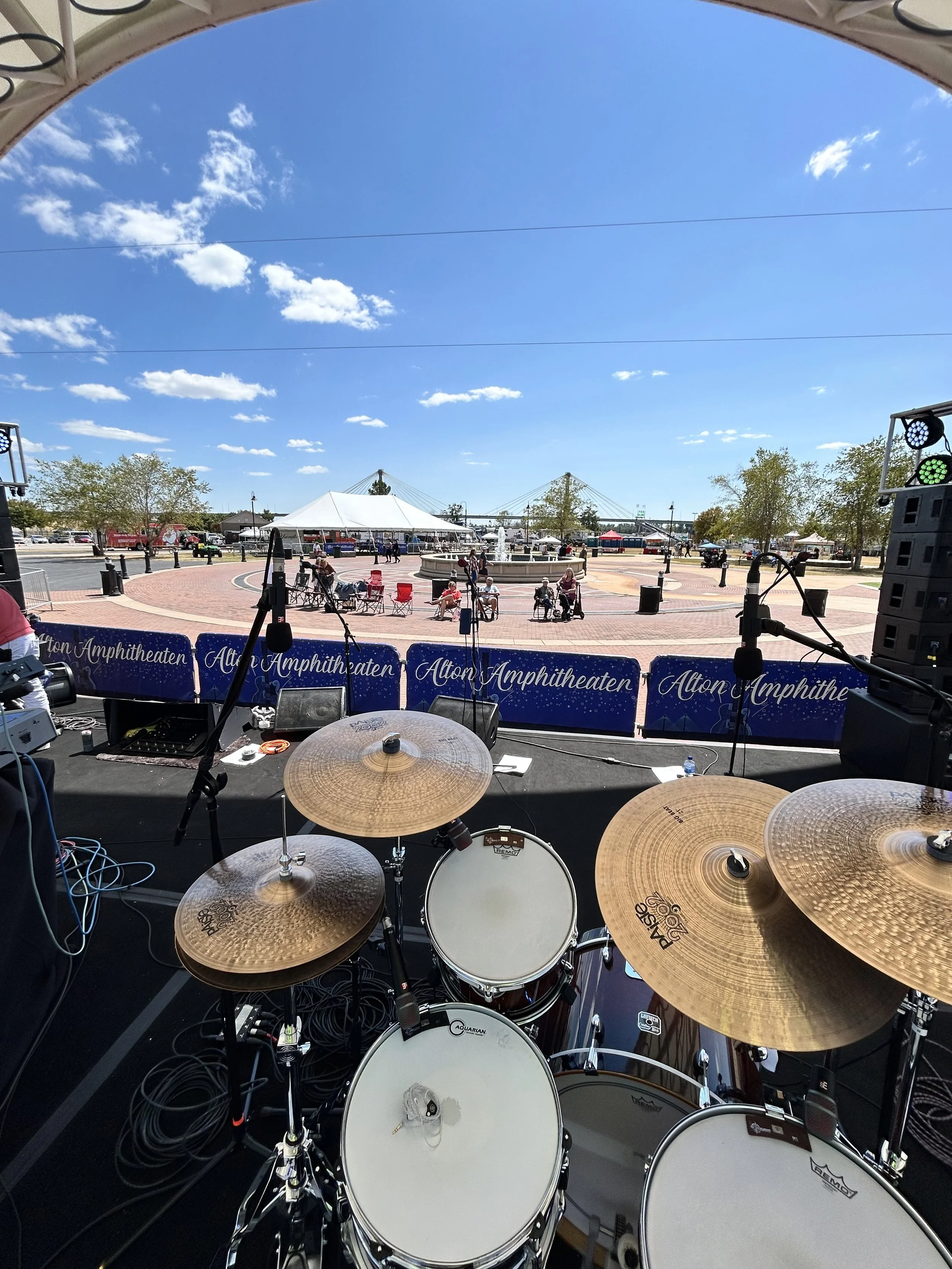 View from stage with drum set, looking out over an outdoor amphitheater with a blue sky, white clouds, trees, a fountain, and people sitting on chairs.