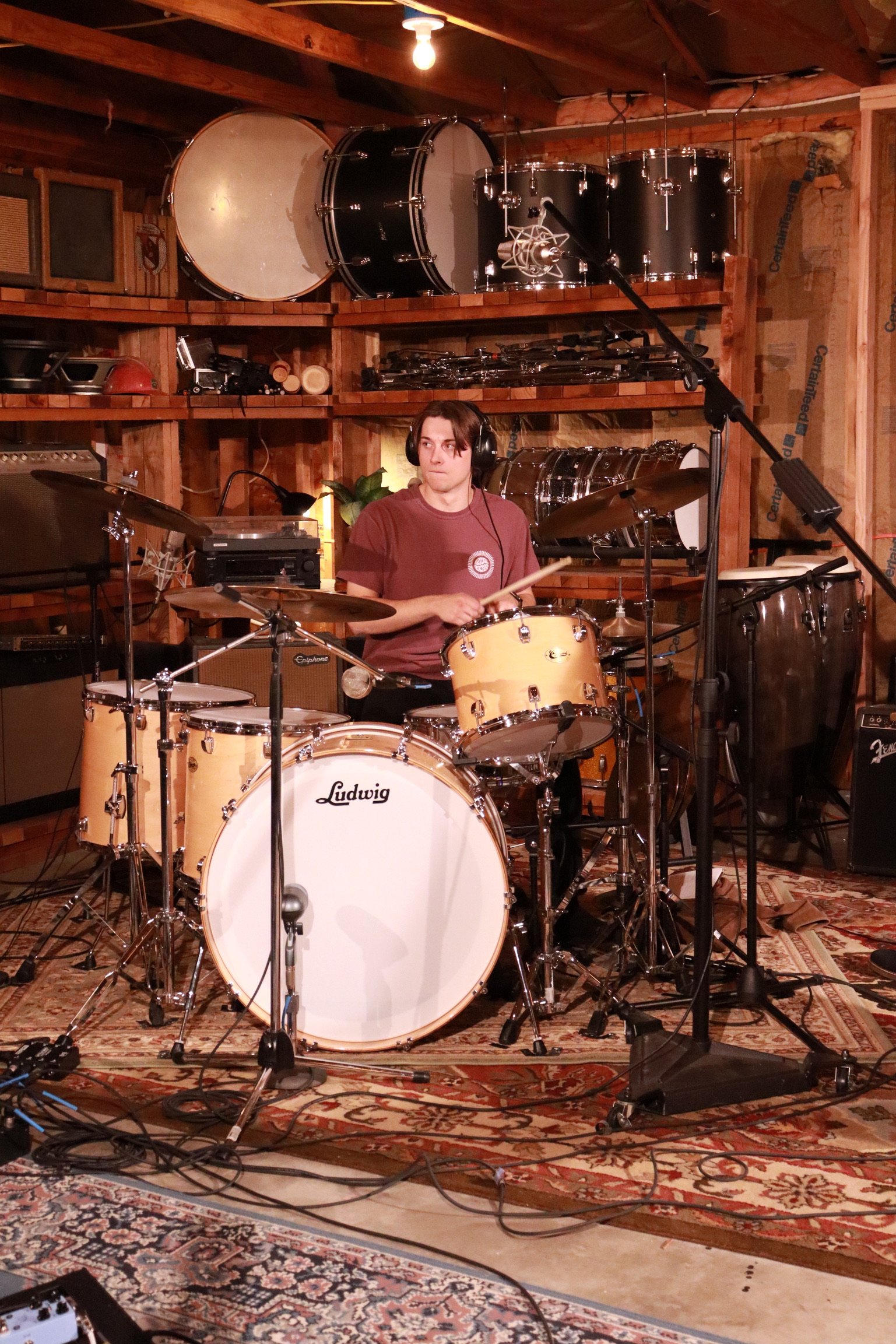 A young man with dark hair wearing a maroon t-shirt is sitting behind a Ludwig drums set in a rustic recording studio, surrounded by various musical instruments and equipment, with drums on shelves behind him and a microphone stand in front.