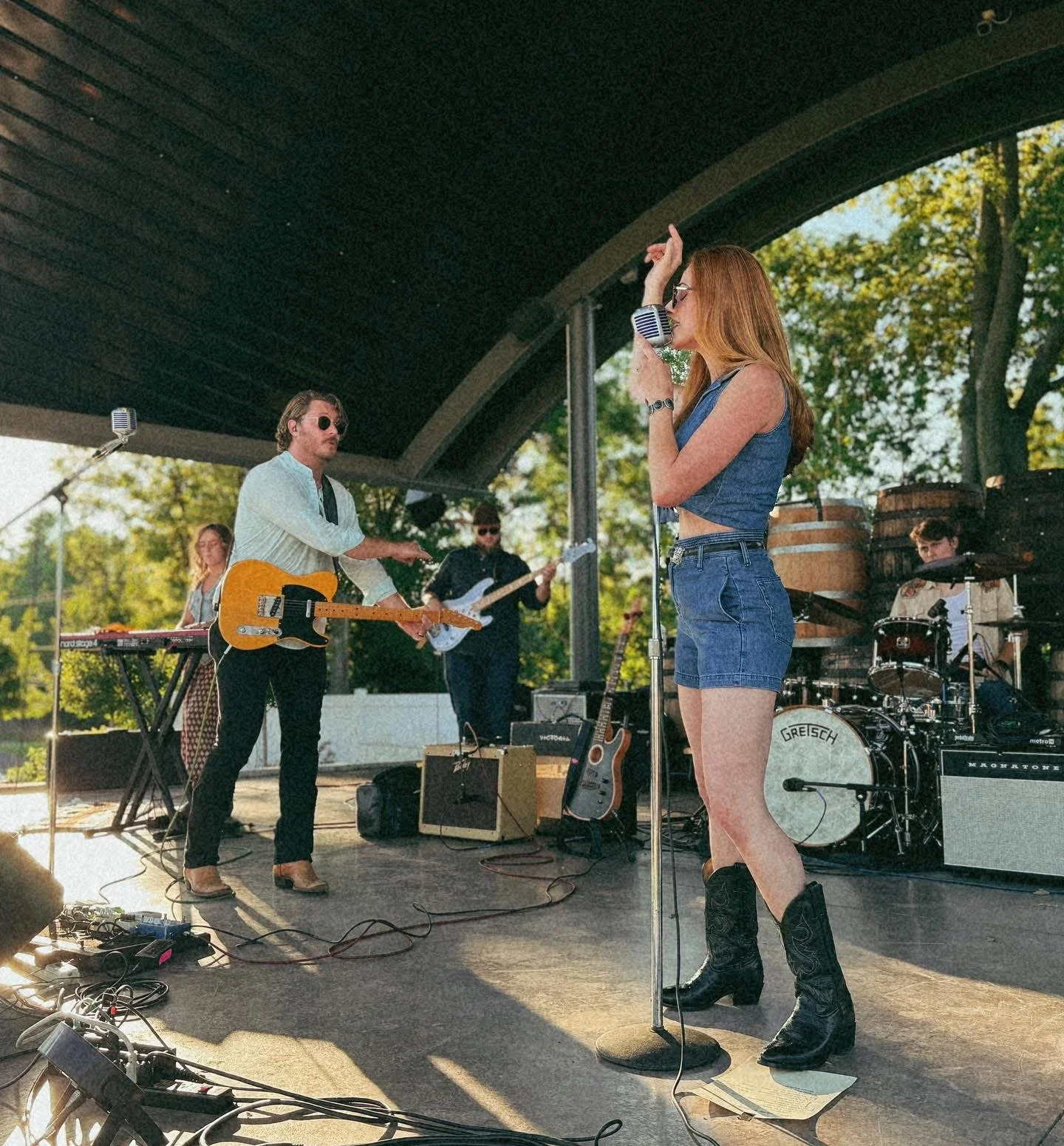 Female singer performing with a band on a stage outdoors during daytime, surrounded by trees.