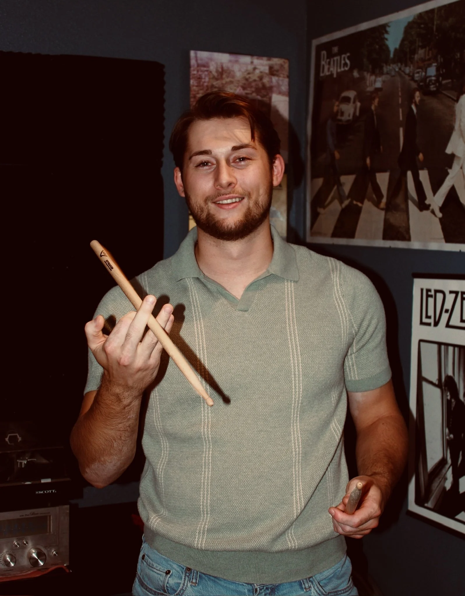 A young man with a beard and short hair smiling at the camera, holding a drumstick in his right hand and a stone in his left hand, standing in a room decorated with Beatles posters.