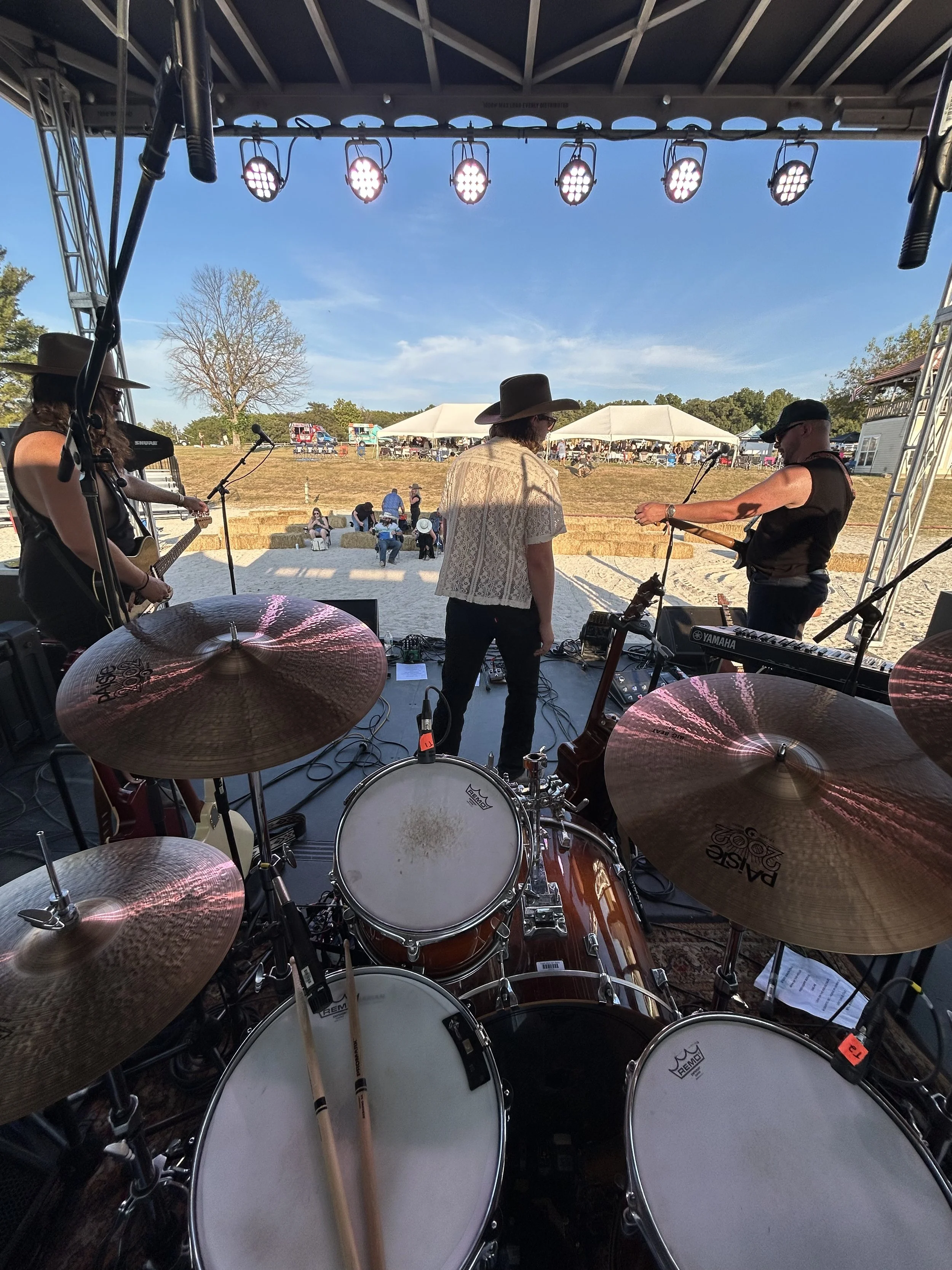 View from stage of a small outdoor country music concert on a sunny day with band members preparing to play. Audience seated on hay bales and in tents in the background.