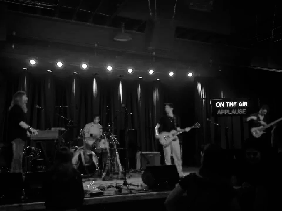 A band performing live on stage in a dimly lit venue, with a drummer, guitarist, and keyboardist, and a sign reading 'ON THE AIR APPLAUSE' in the background.