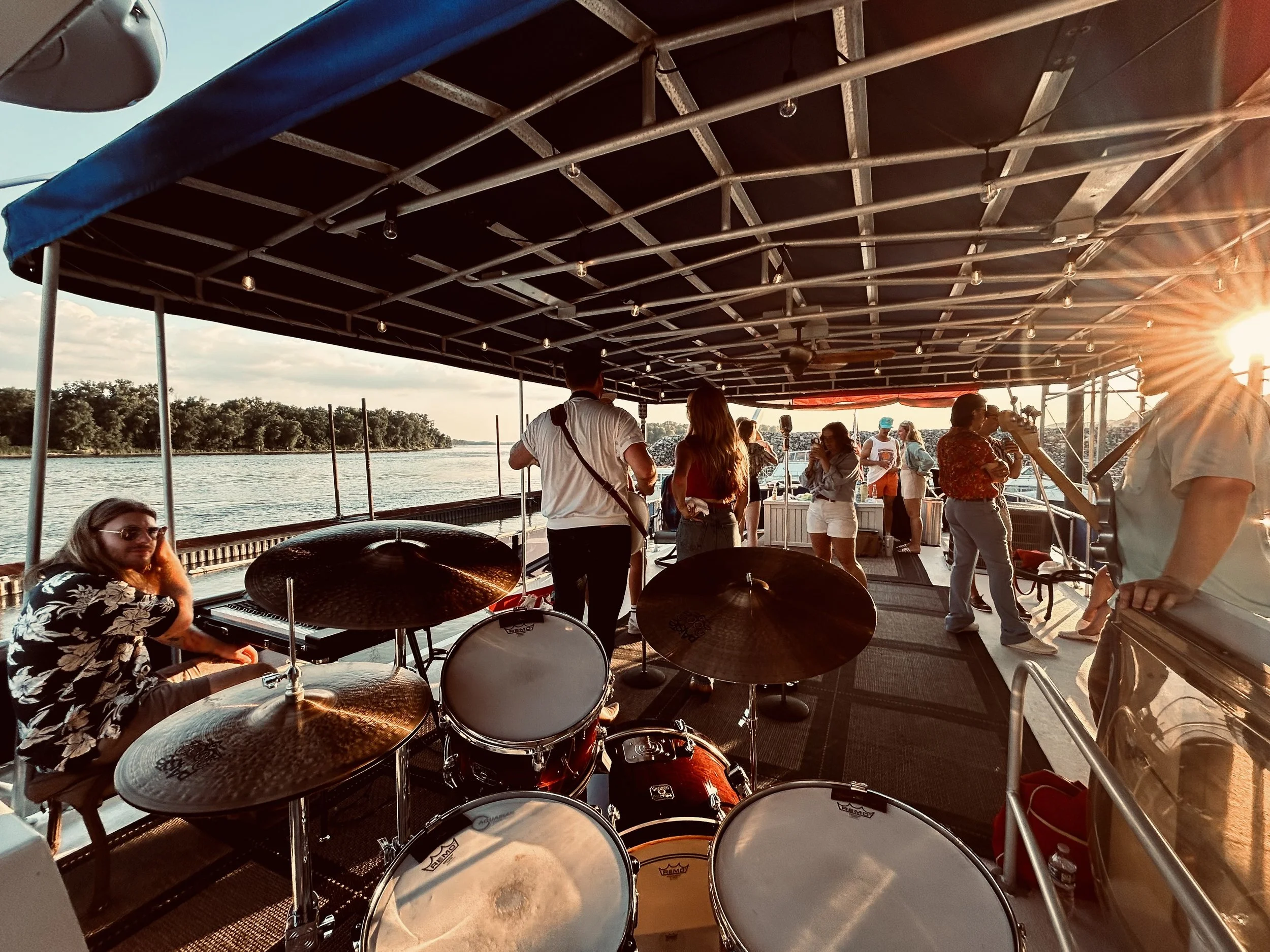 People enjoying a boat cruise on a river at sunset, with a drum set and window on the boat's deck.