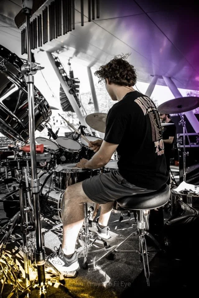 A person playing electronic drums on stage with a keyboard above, during a live performance.