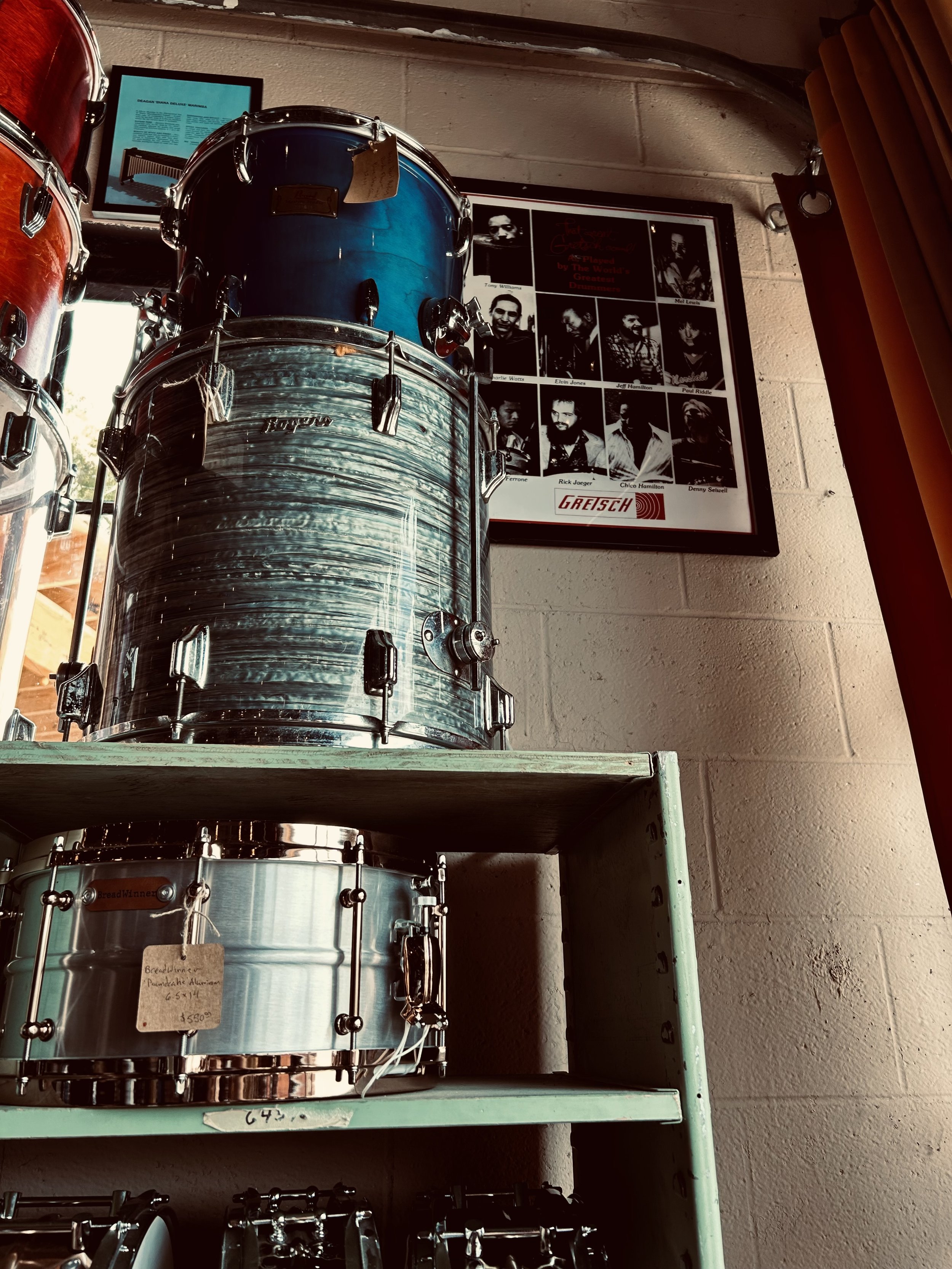 Display of drum kits on a shelf, with a framed black-and-white band photograph and a promotional poster on a brick wall in the background.