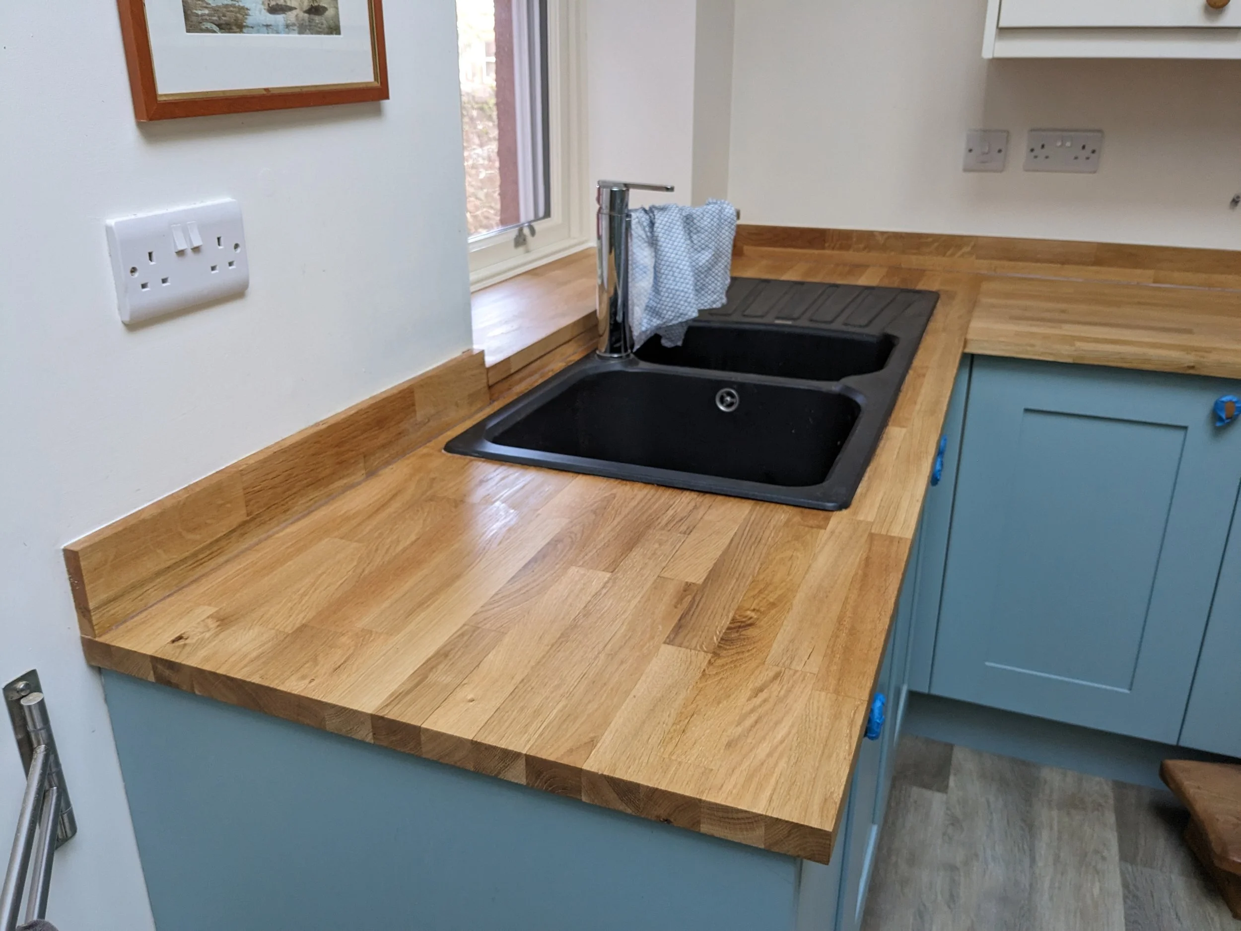 Kitchen countertop with a black double sink and a small towel hanging from the faucet, next to a window and a wooden backsplash.