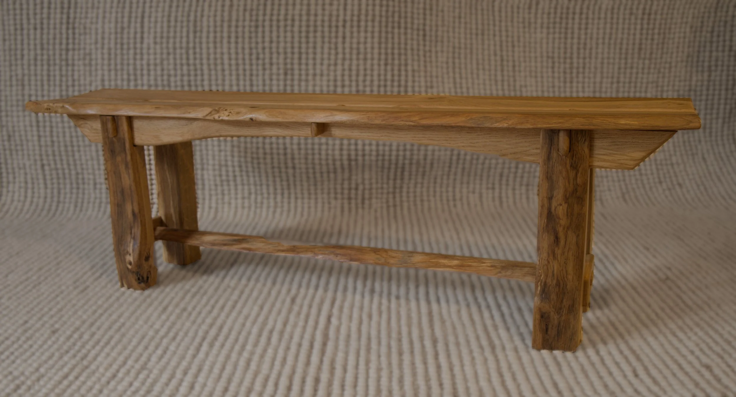 A small handcrafted wooden bench with a simple design, placed on a textured beige fabric background.