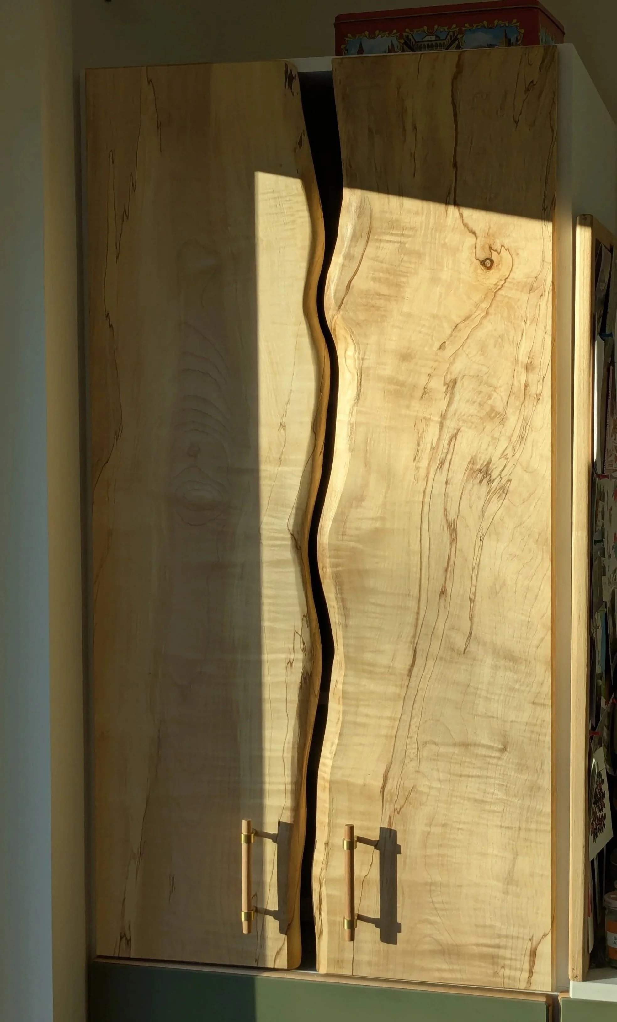 A wooden cabinet with a unique, wavy-open front design and two brass handles in a room with a bookshelf and a decorative tin on top.