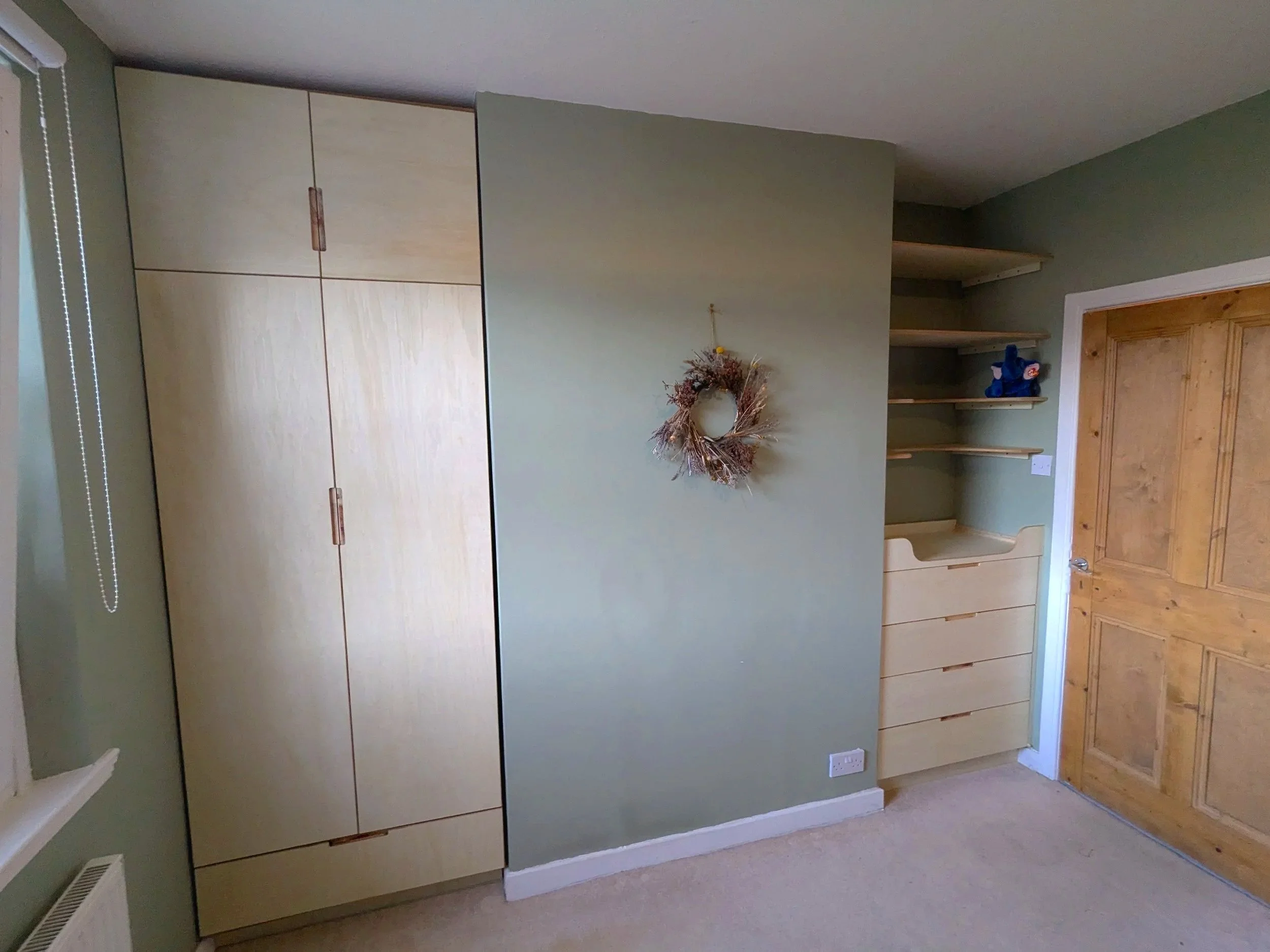 A room with light green walls, a wooden door, built-in wooden shelves, and a light-colored wooden wardrobe. A wreath hangs on the wall beside the shelves.