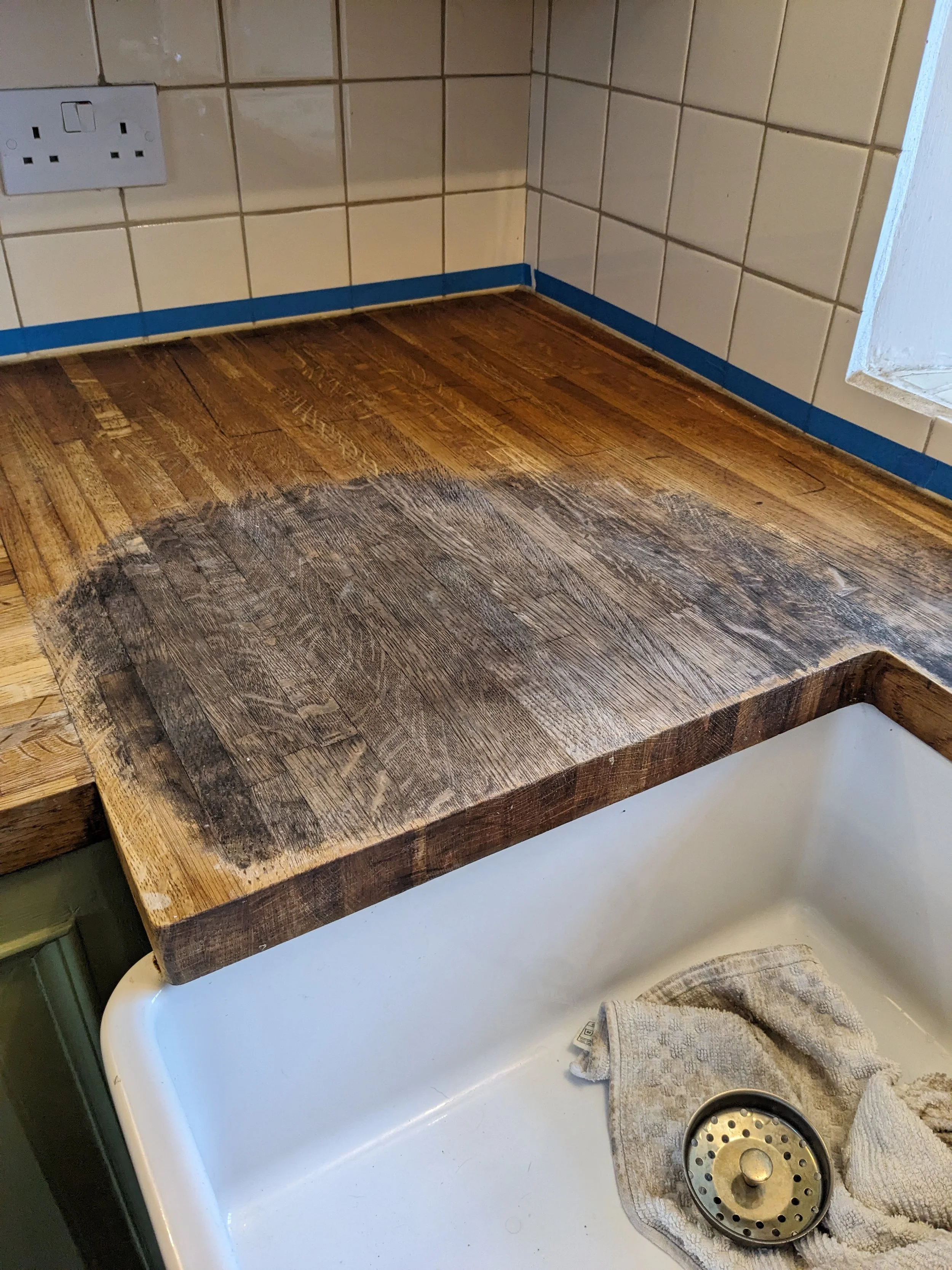 A wooden kitchen countertop over a white sink, with a towel and sink drain inside the sink, and tiled walls with an electrical outlet in the background.