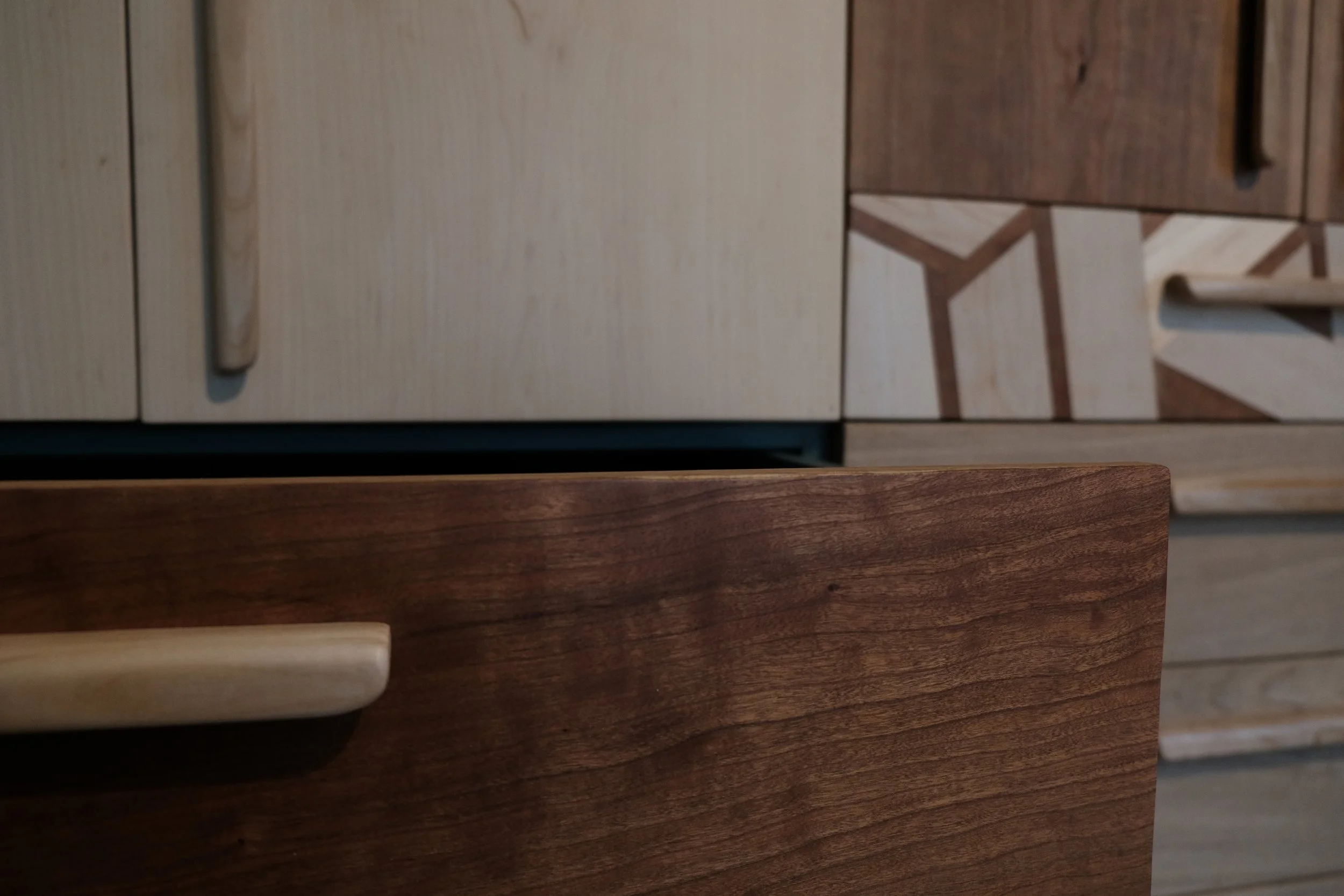 Close-up of a wooden table surface with a light-colored handle on the left, with background showing a cabinet with mixed wood tones and geometric designs.