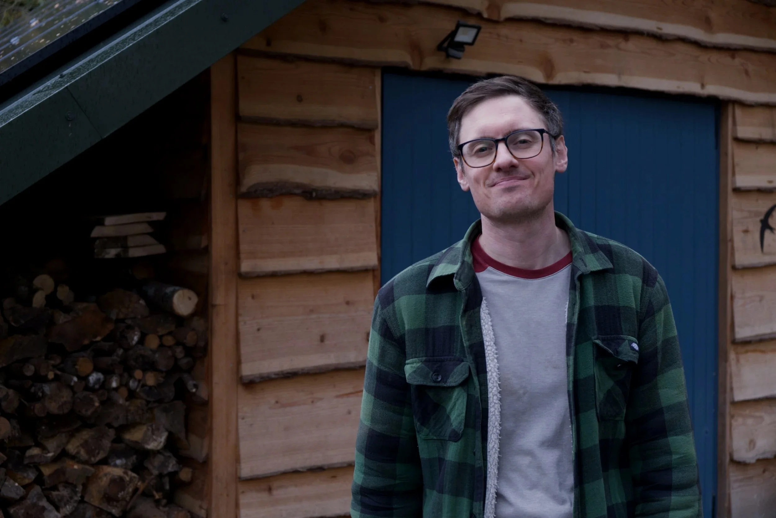 A man with glasses and short dark hair standing outside a wooden shed, smiling at the camera.