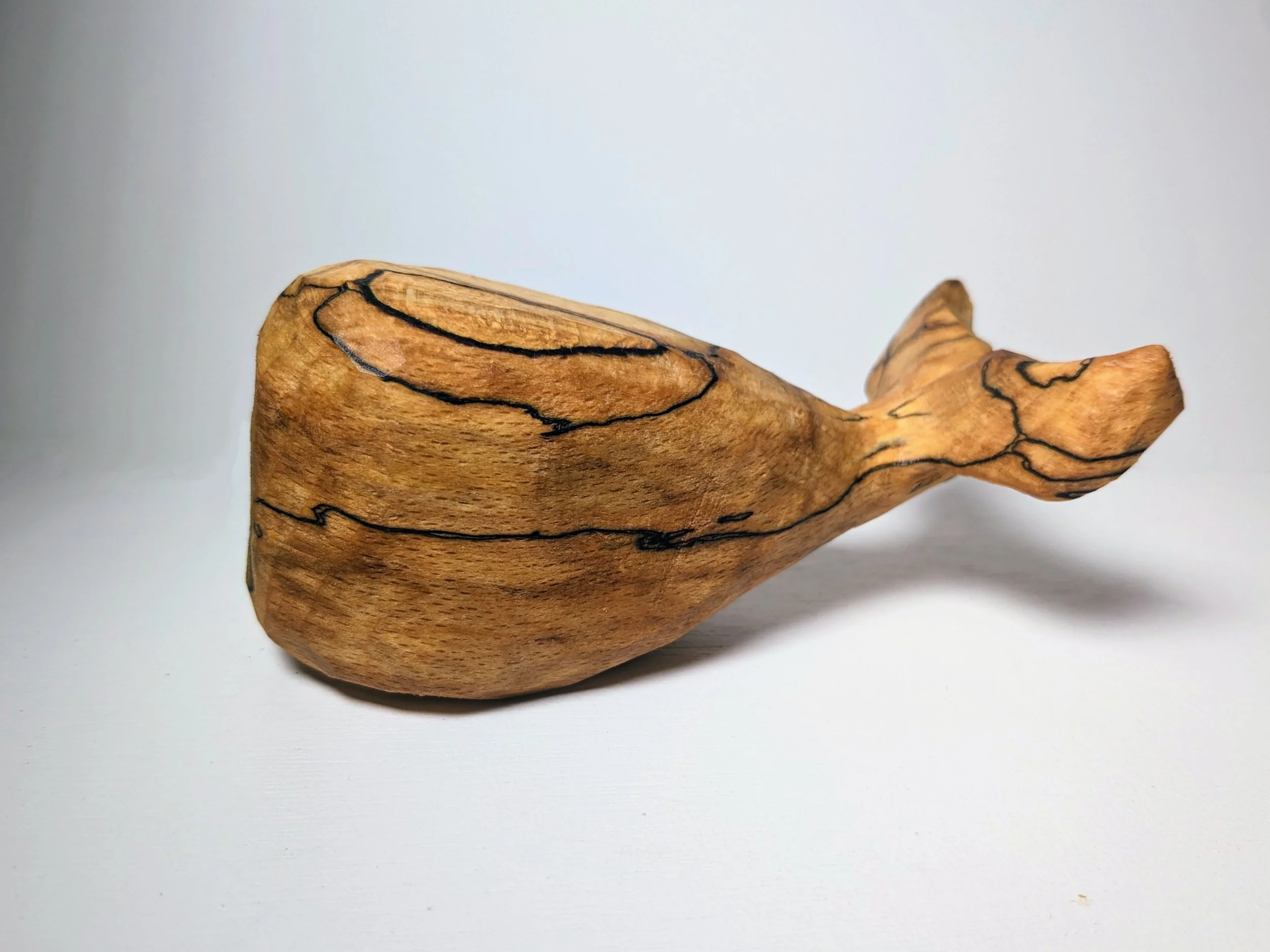 A wooden sculpture resembling a whale, made from a single piece of wood with natural grain and dark lines.