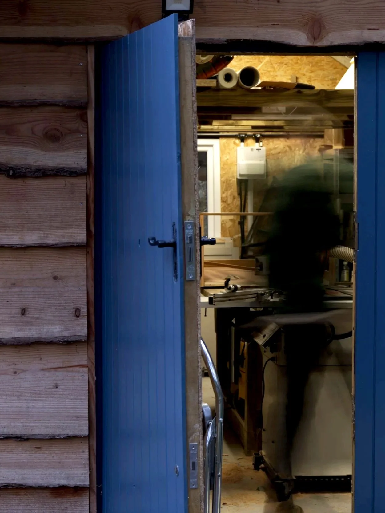 View through an open blue door into a woodworking workshop with various tools and materials.