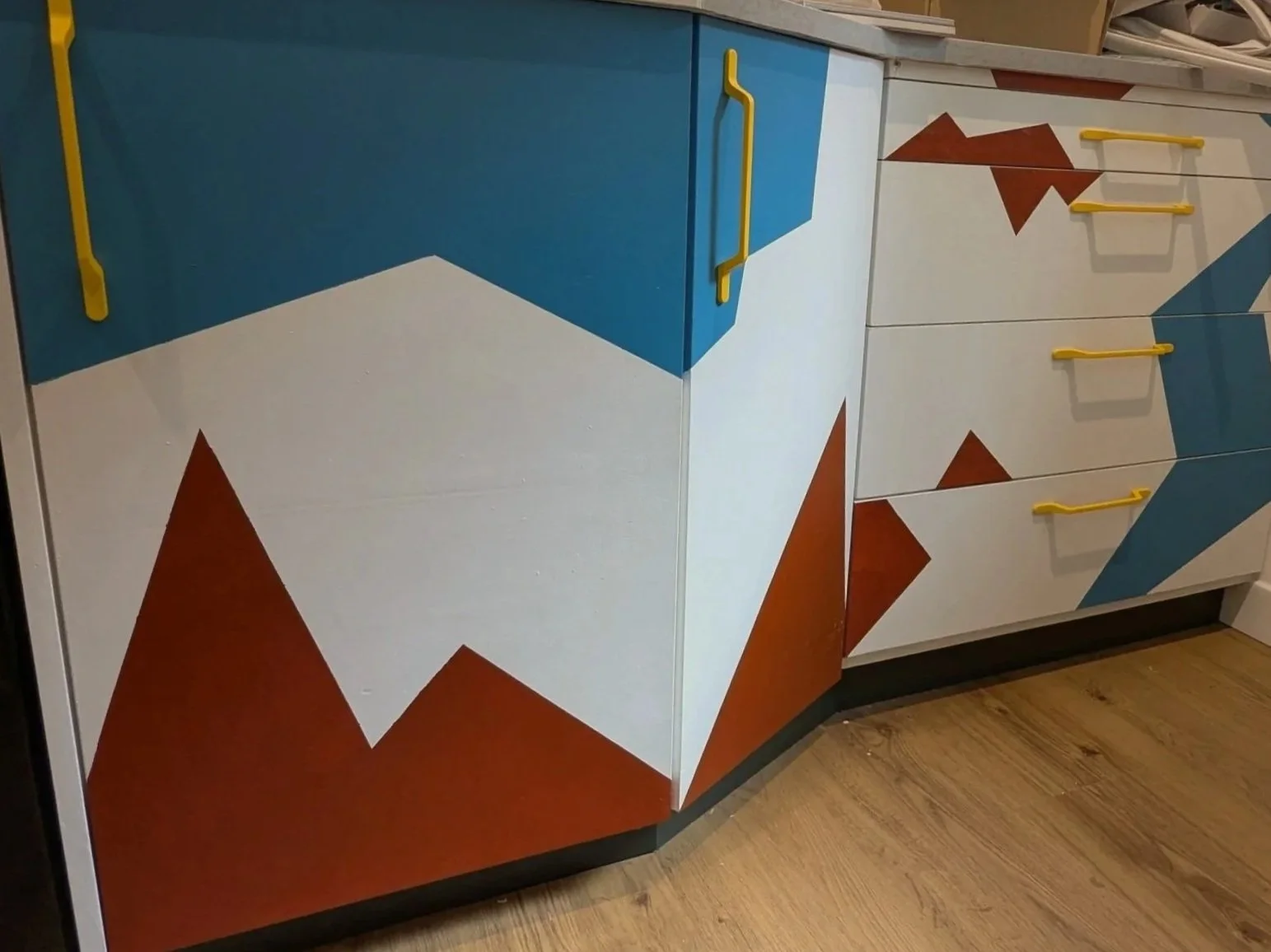 Colorful kitchen cabinet with a geometric mountain design painted on the front, featuring white, blue, and red triangles, and yellow handles.