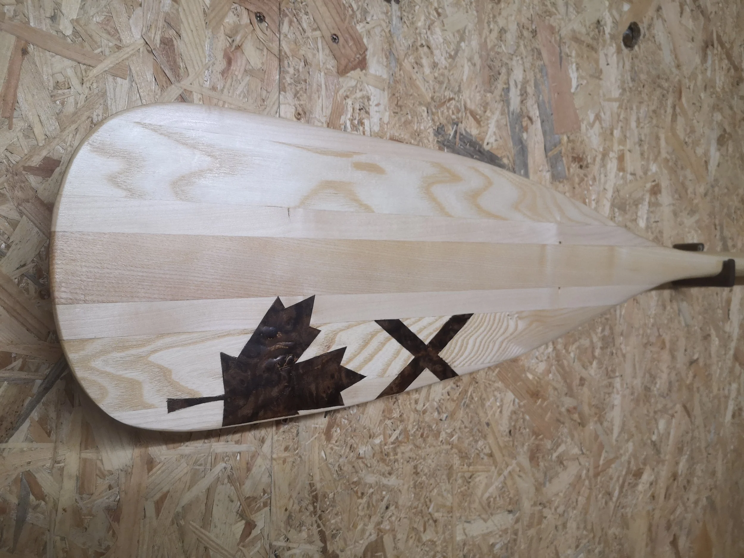 Wooden boat with a maple leaf and 'X' painted in dark color on the side, resting on a plywood floor.
