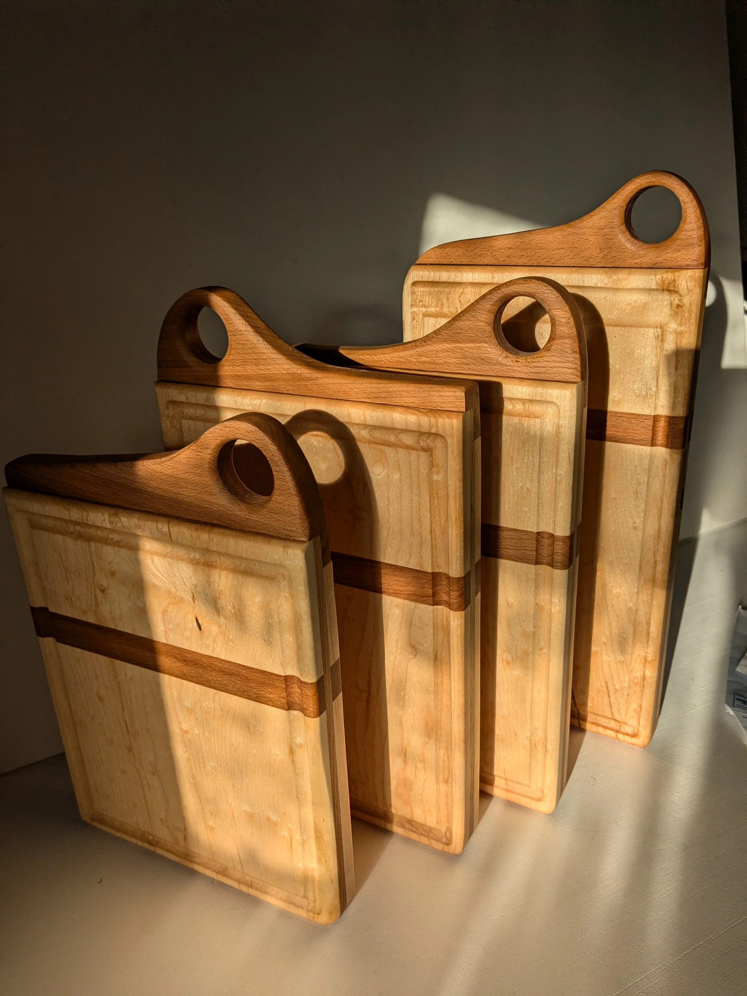 Three wooden cutting boards of varying sizes, arranged in a row, with sunlight casting shadows on a grey wall background.