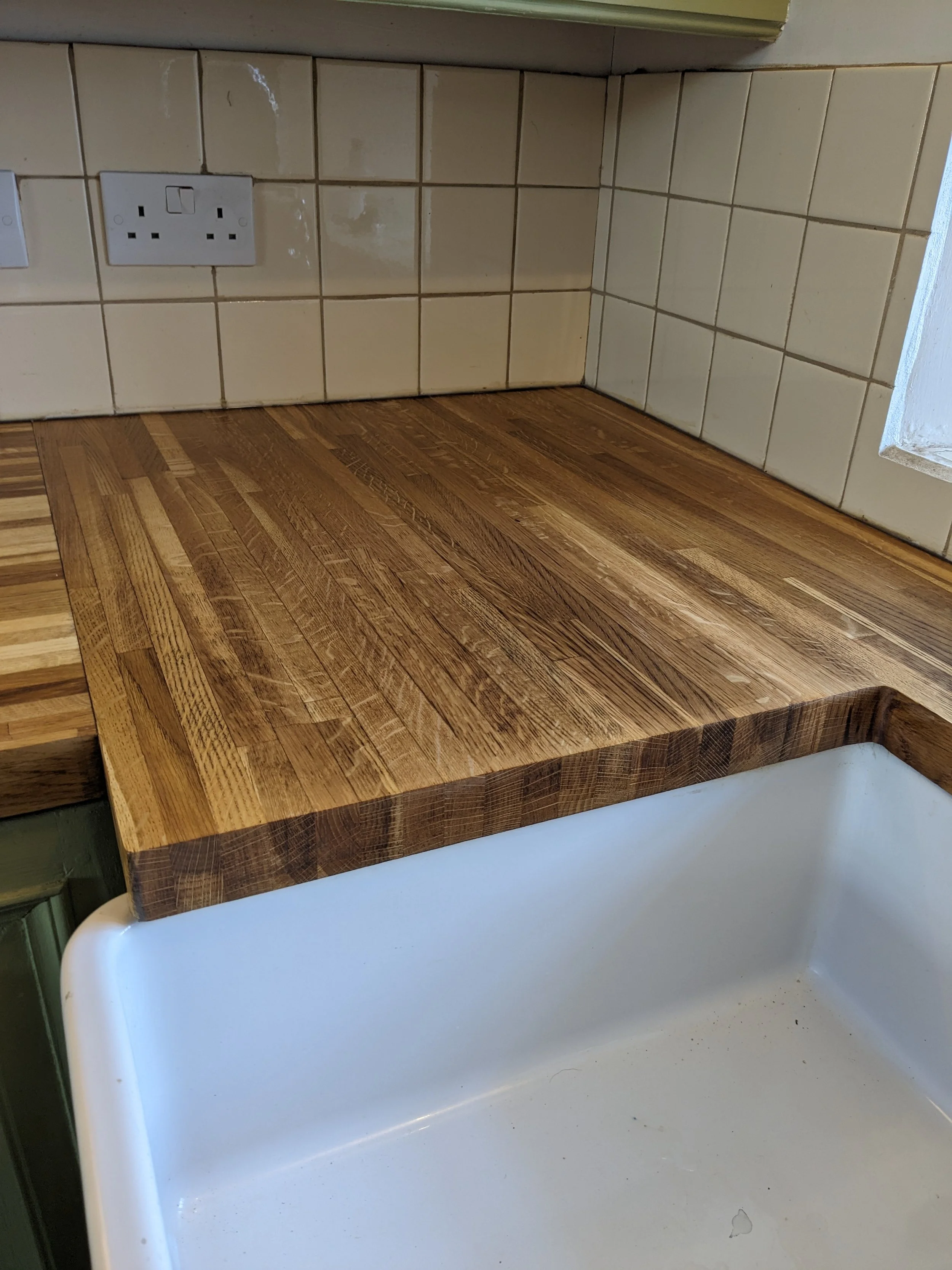 Wooden kitchen countertop next to a white sink with a tiled wall and electrical outlets in the background.