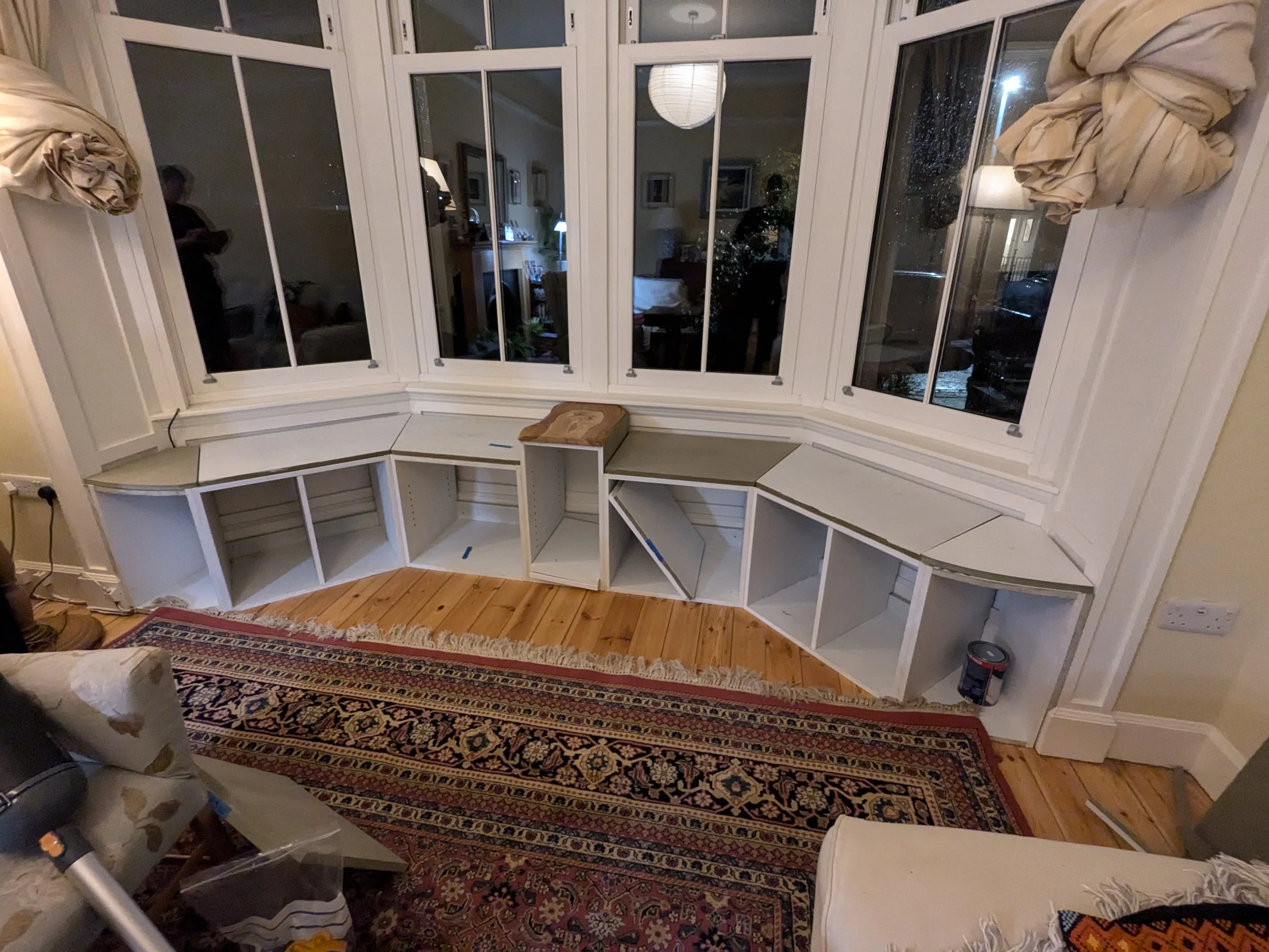 Living room with large bay window, built-in white bookcase shelves, and a detailed patterned rug on hardwood floor. The shelves are partially assembled with some empty compartments and one with a small wooden piece on top.