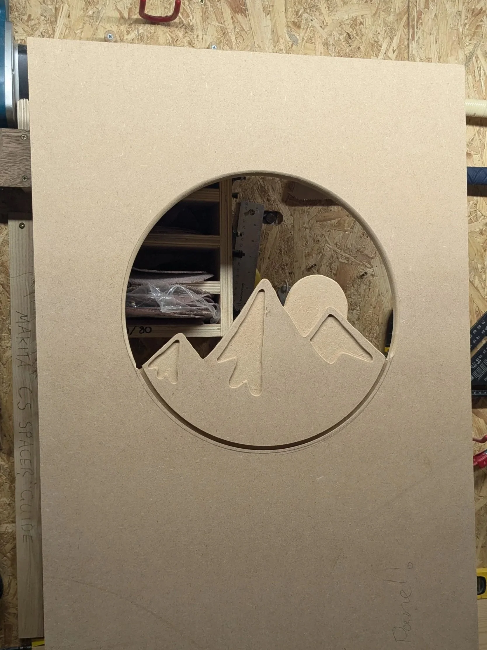 Wooden panel with a circular cutout depicting mountain peaks and a sun or moon, in a workshop setting.