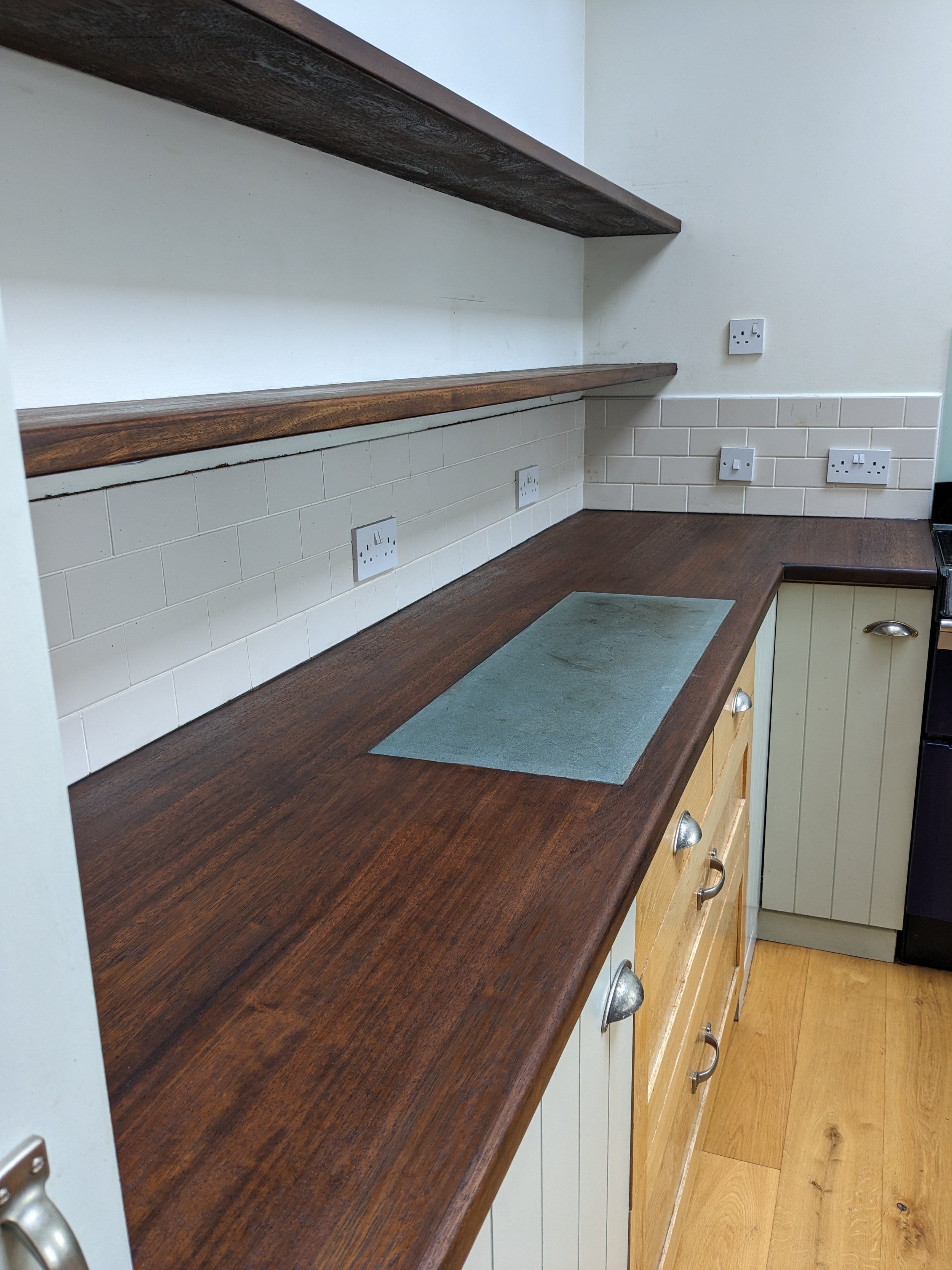 A kitchen countertop with a dark wooden surface, beige kitchen cabinets with metallic handles, and cream-colored tile backsplash. There are multiple electrical outlets along the wall above the counter and shelves.