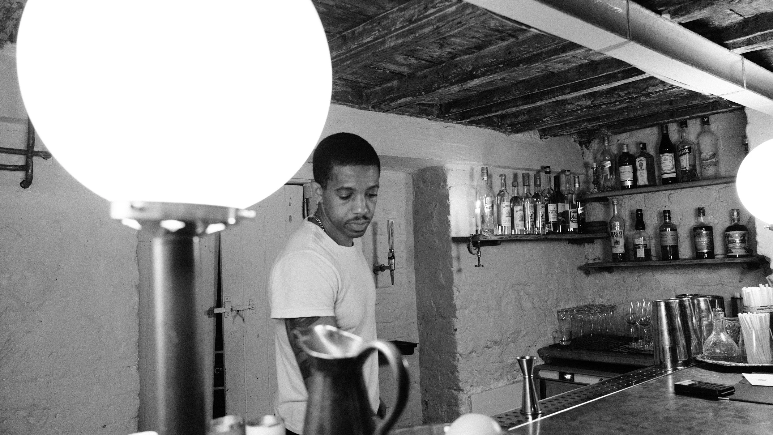 A man behind a bar counter in a dimly lit room with shelves of liquor bottles in the background. Large glowing spherical lamps are hanging from the ceiling.