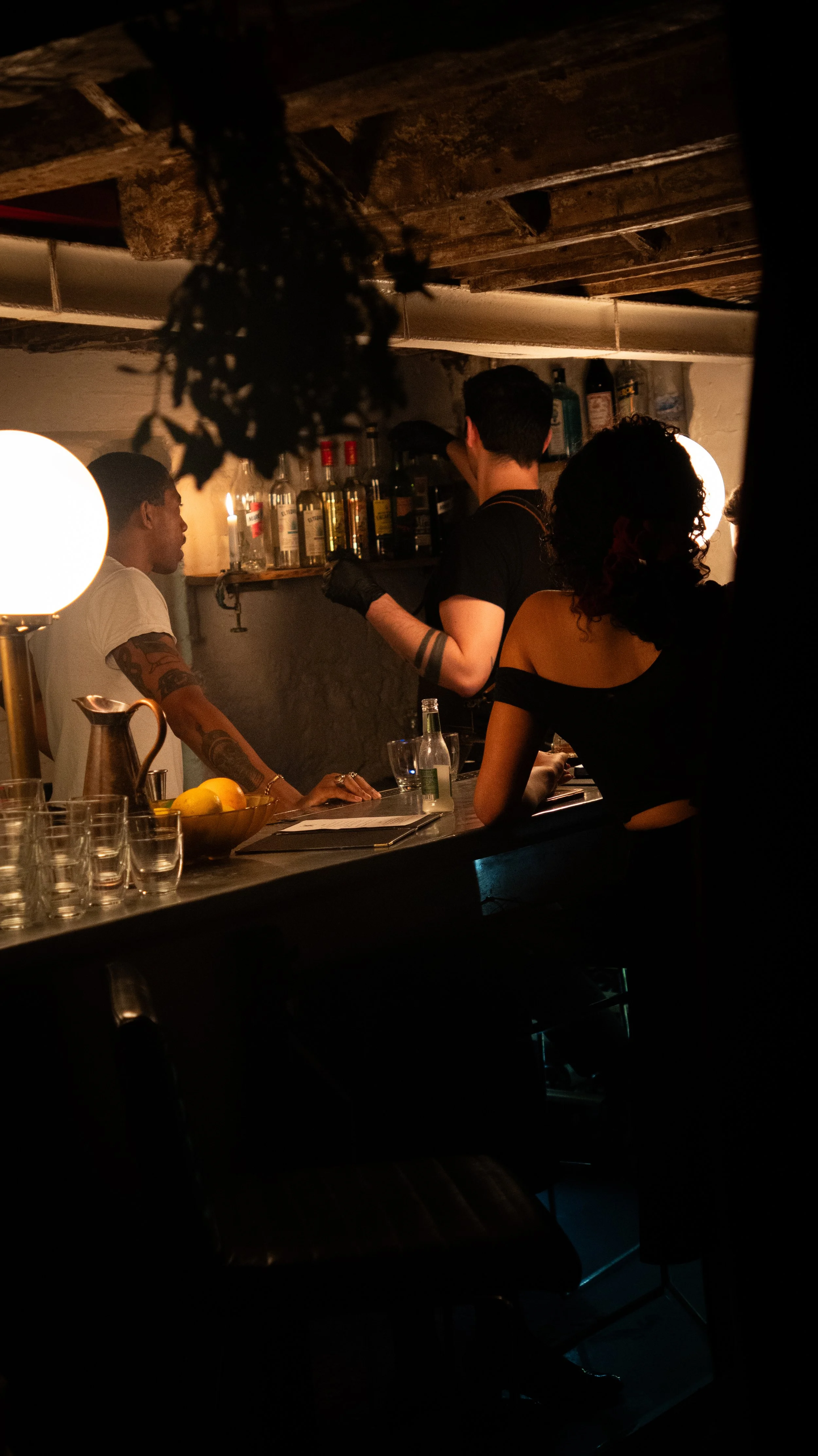 Three people working behind a bar in a dimly lit setting, with bottles on a shelf and glasses on the counter.