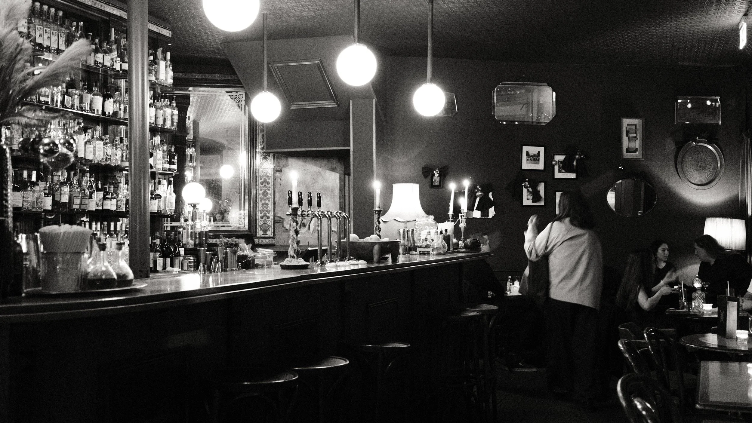 A black-and-white photo of a dimly lit bar with shelves of liquor bottles, a counter with beer taps, and a few patrons sitting and conversing.